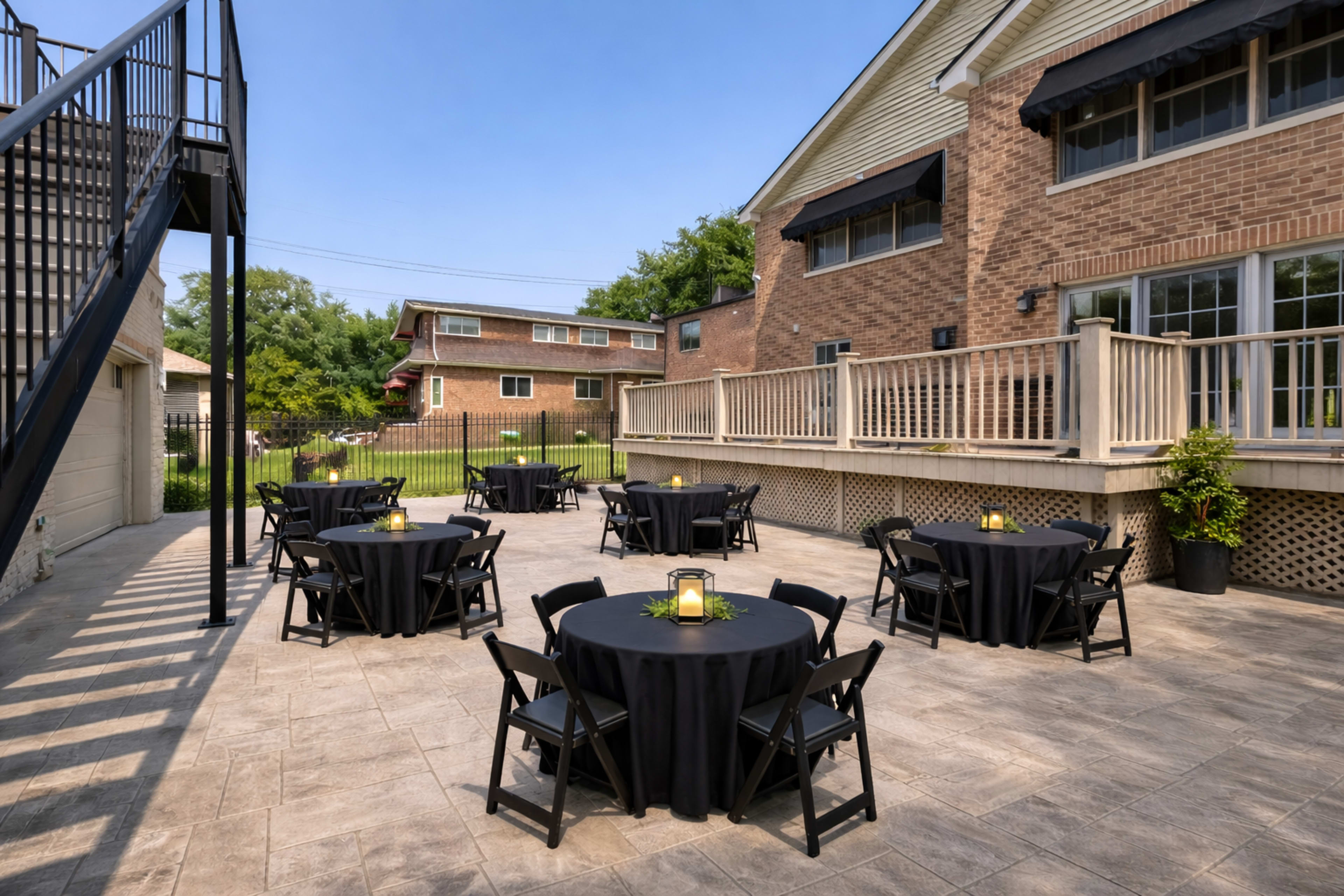 The image shows an outdoor patio area with several round tables covered in black tablecloths, surrounded by a brick building and greenery.