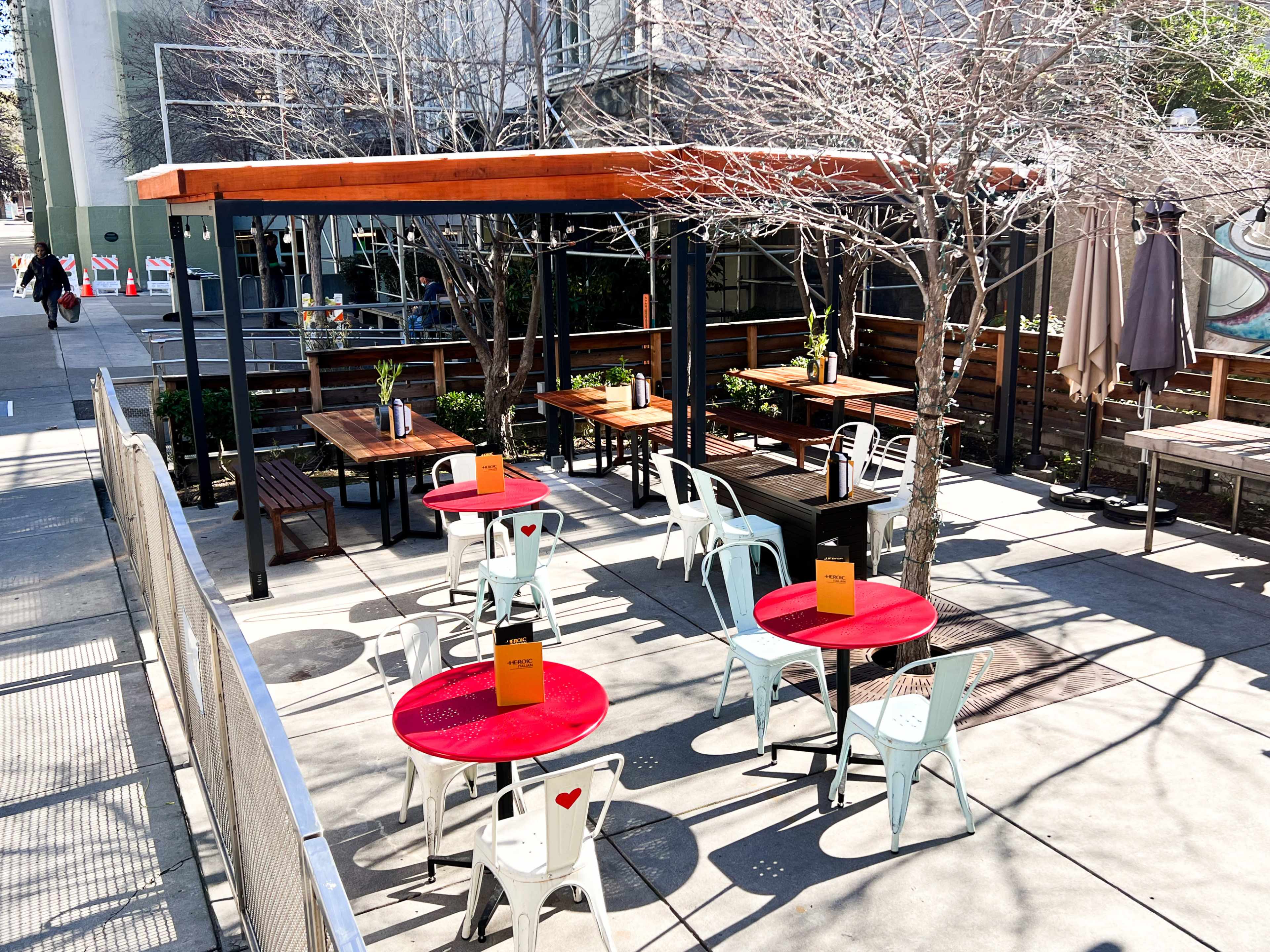 An outdoor dining area features several red tables surrounded by white chairs, with a shaded canopy and trees in the background.