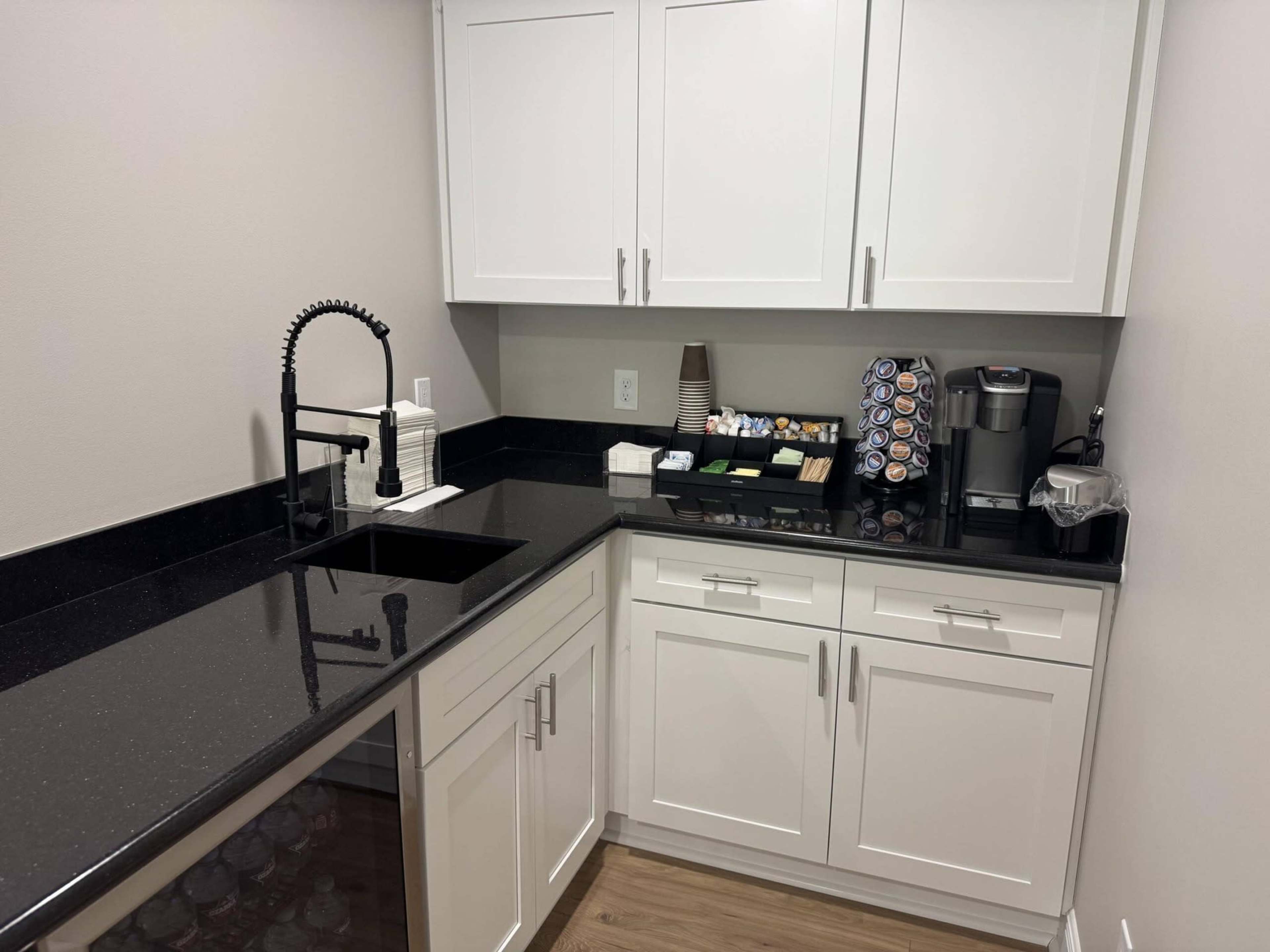 The image shows a modern kitchenette with white cabinets, a black countertop, a sink, and an arrangement of coffee-making supplies.