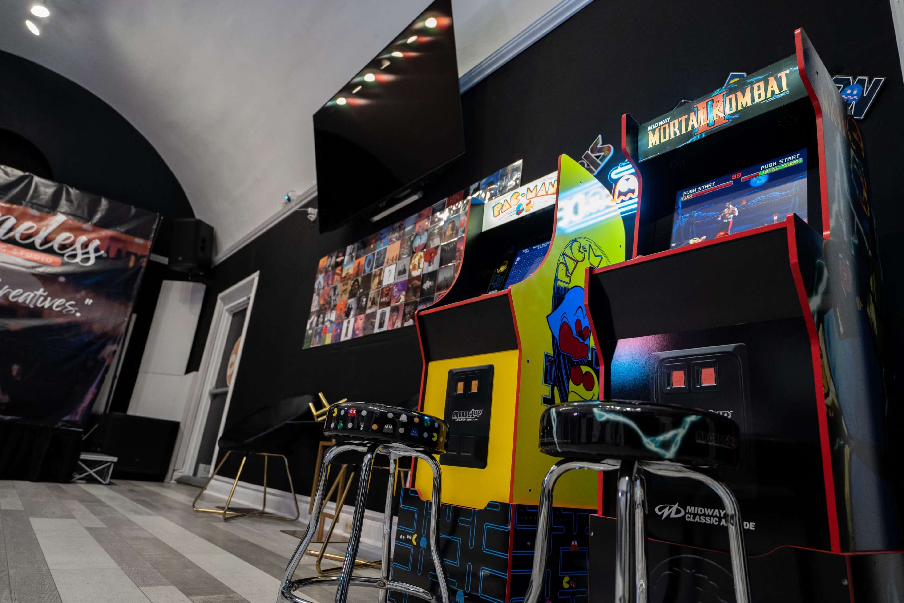 The image shows a game room featuring two vintage arcade machines, "Pac-Man" and "Mortal Kombat," along with bar stools and a wall of framed artwork.