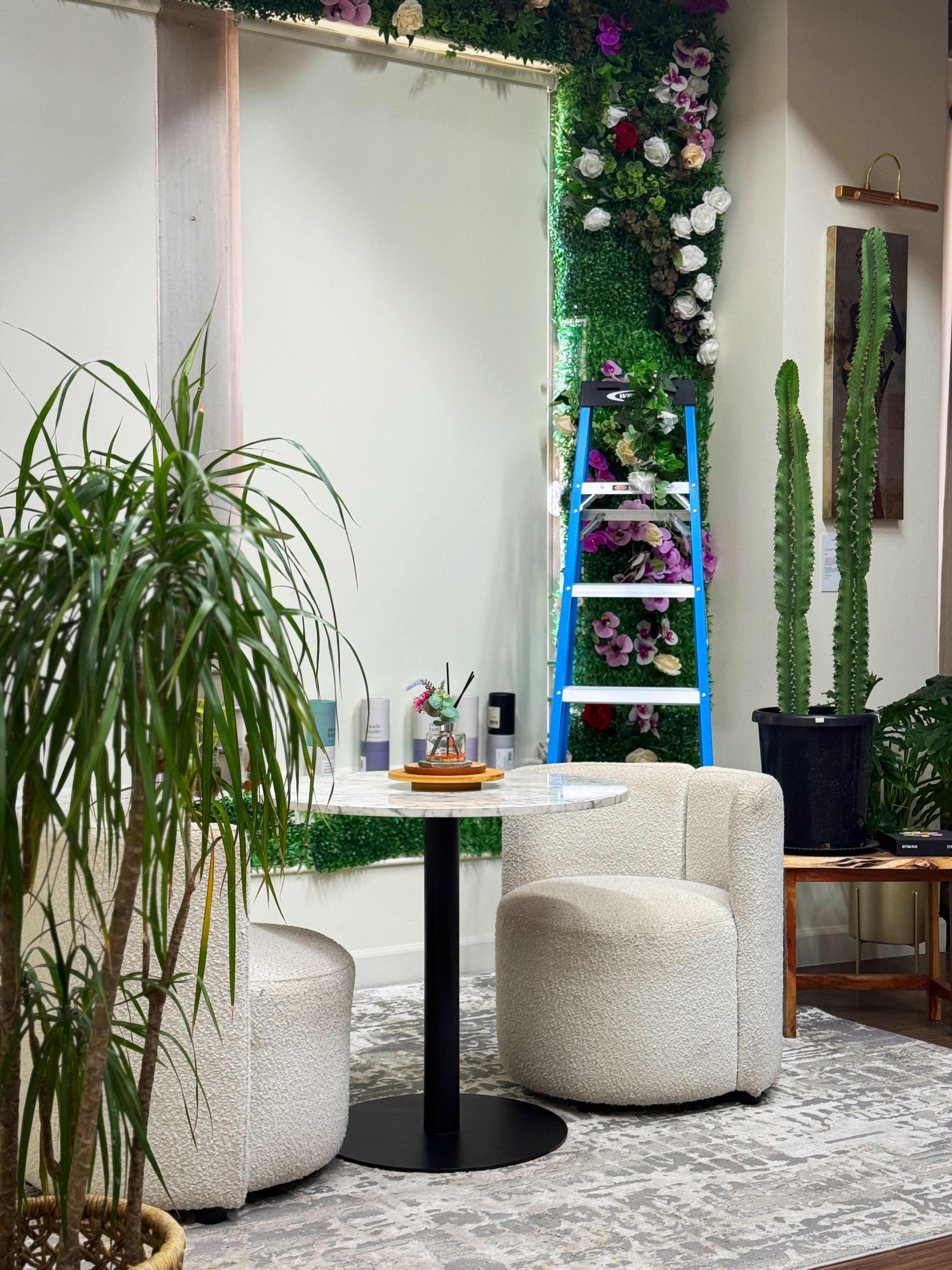 A small interior space with a round table and two upholstered chairs, surrounded by plants, a tall ladder, and a wall adorned with greenery and flowers.