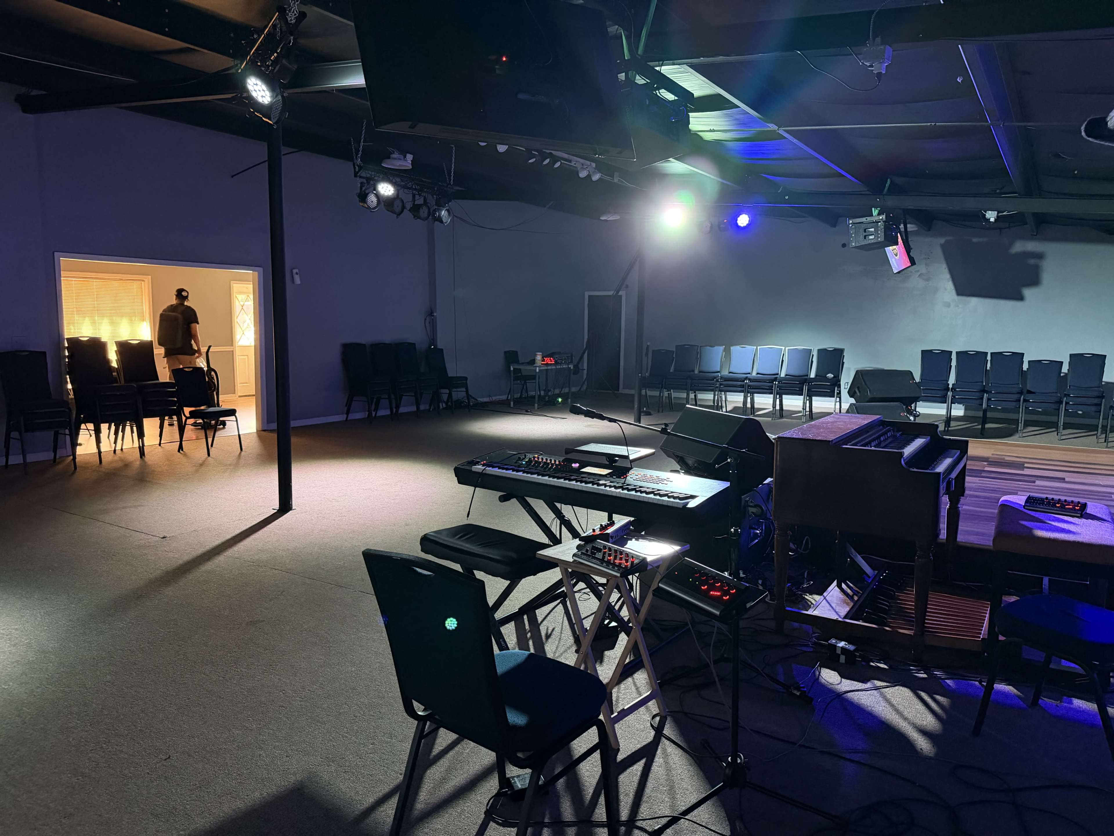 A music performance space is set up with a keyboard and various equipment in the foreground, while chairs are arranged in rows in the background.