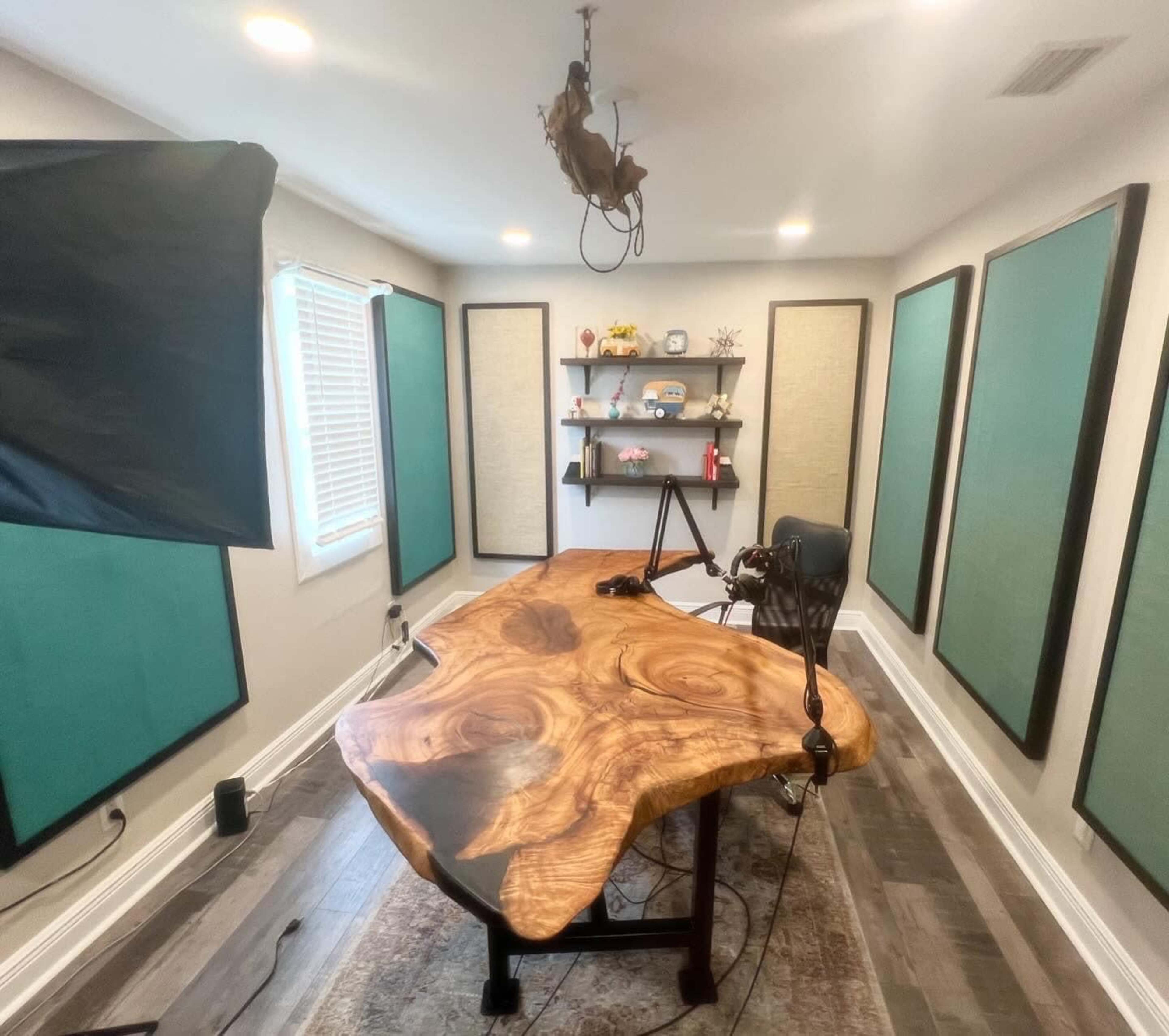 The image shows a modern recording studio featuring a large, natural wood table, a microphone setup, and acoustic panels on the walls.