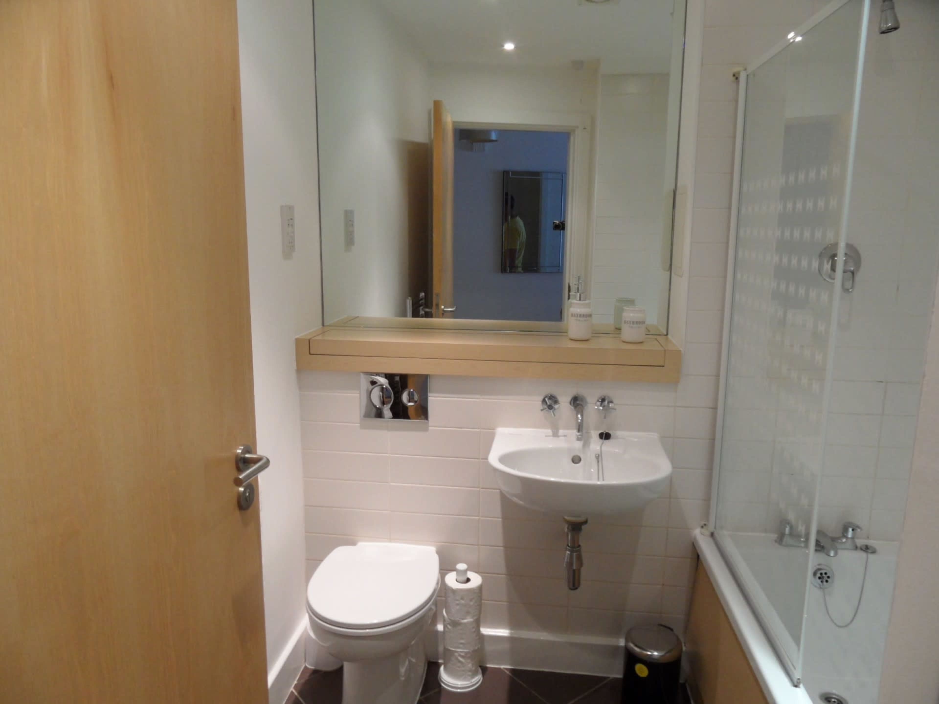 A compact bathroom featuring a toilet, a sink with a mirror above, and a bathtub with a glass shower screen.