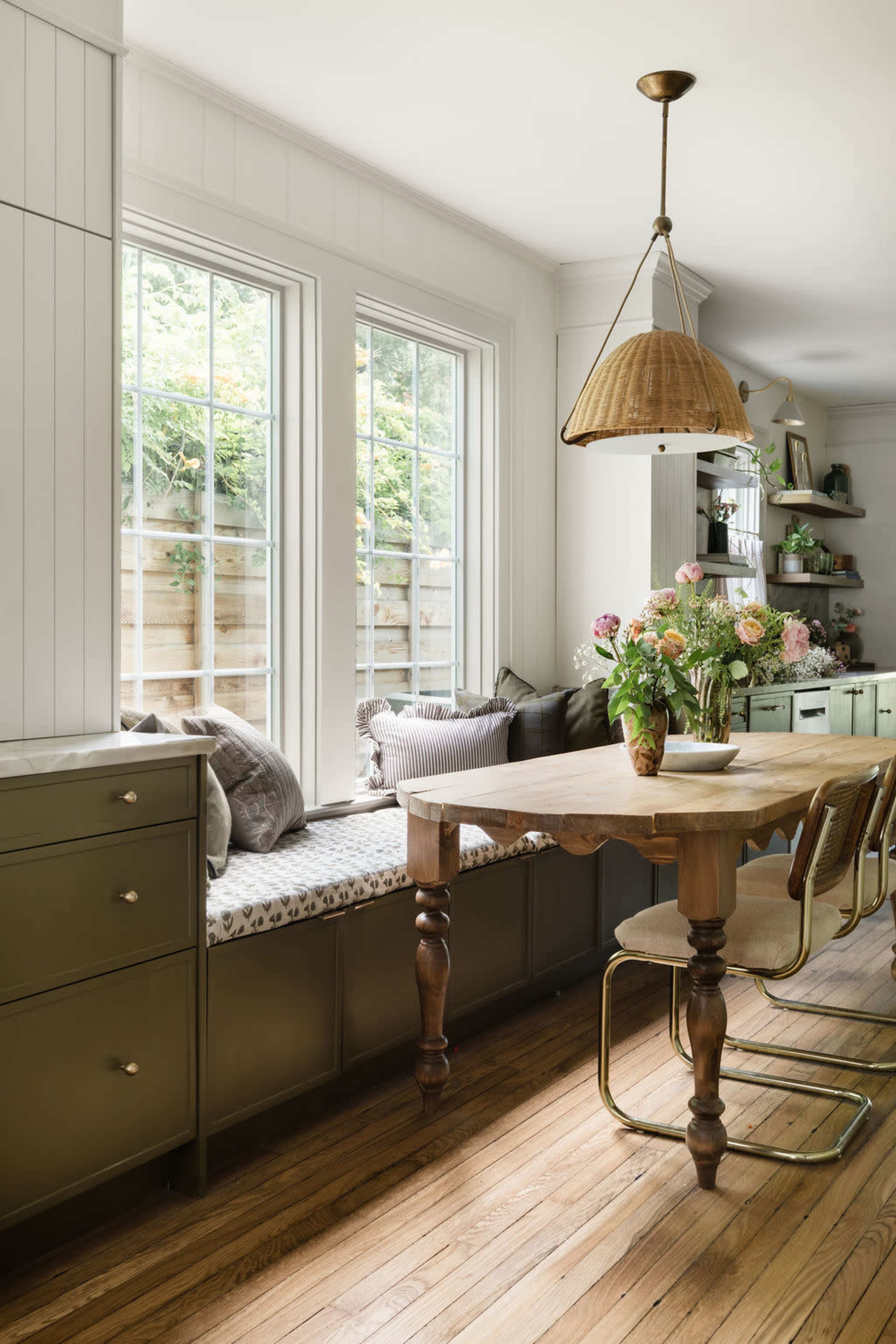 The image shows a dining area with a wooden table surrounded by light-colored chairs, a window seat adorned with cushions, and a vase of flowers on the table.