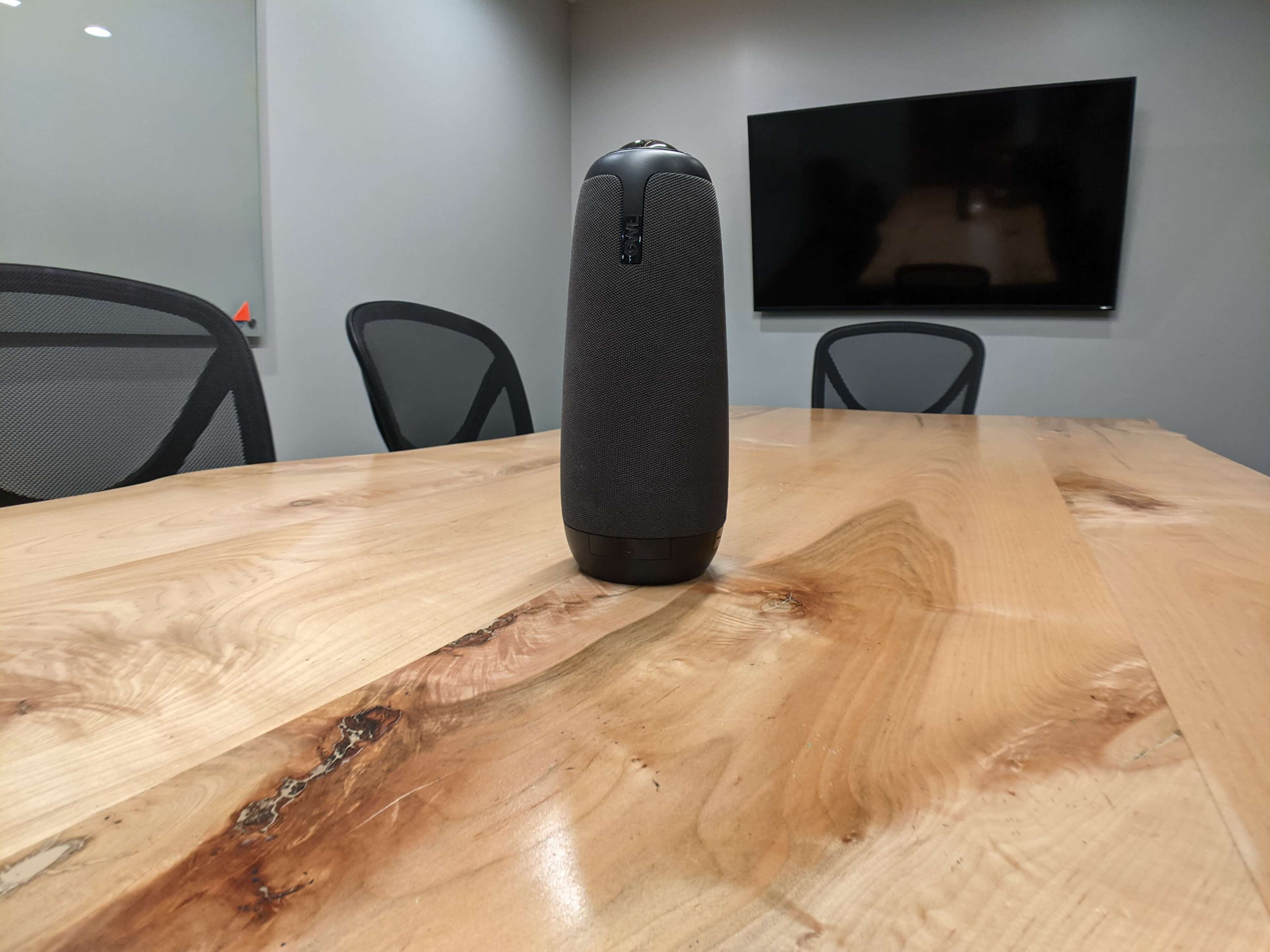 A Bluetooth speaker stands on a wooden conference table in a meeting room with a large screen in the background.