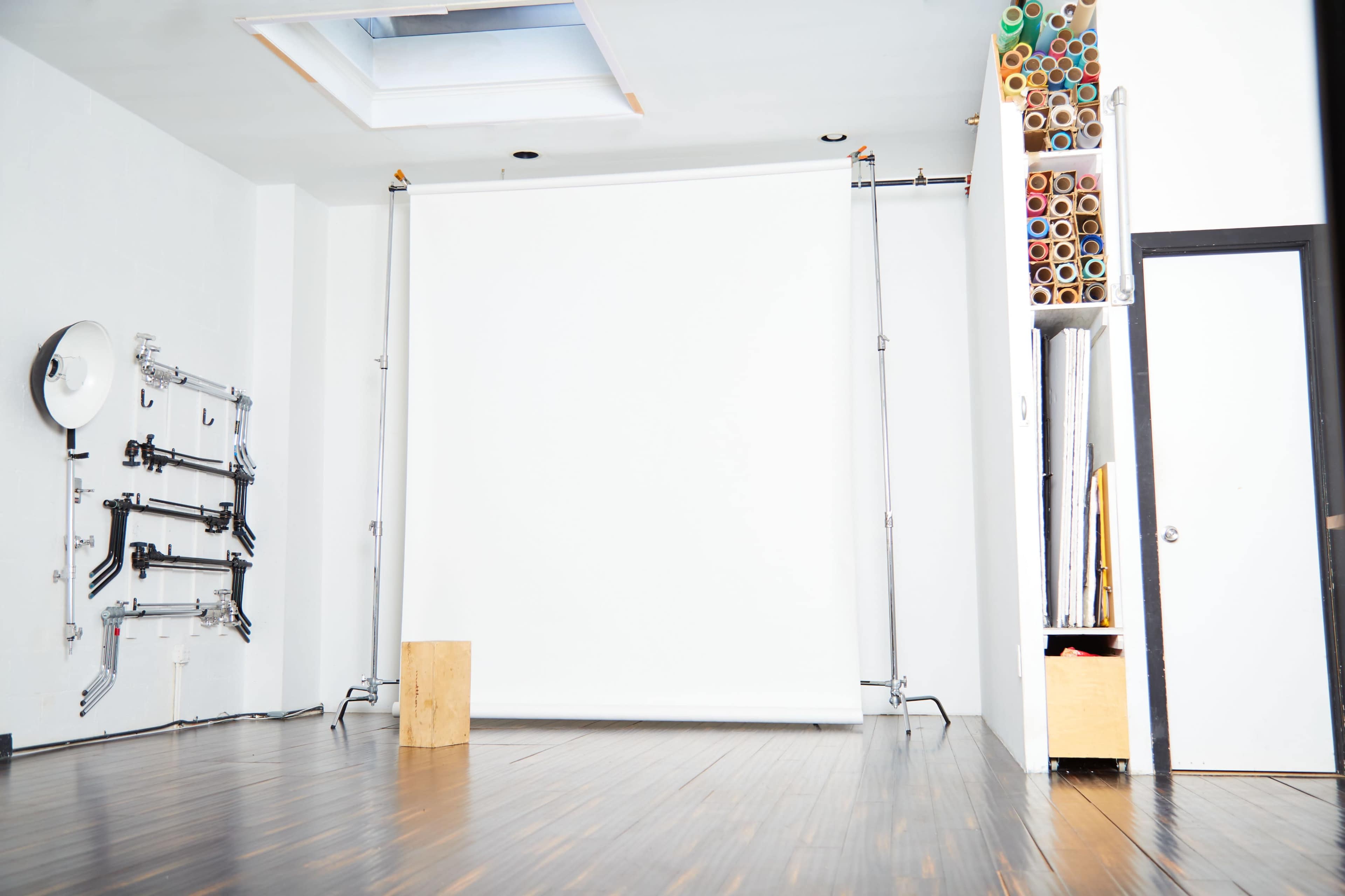 The image shows a bright, empty studio space featuring a white backdrop supported by metal stands, wooden flooring, and a shelving unit filled with rolls of colorful paper.