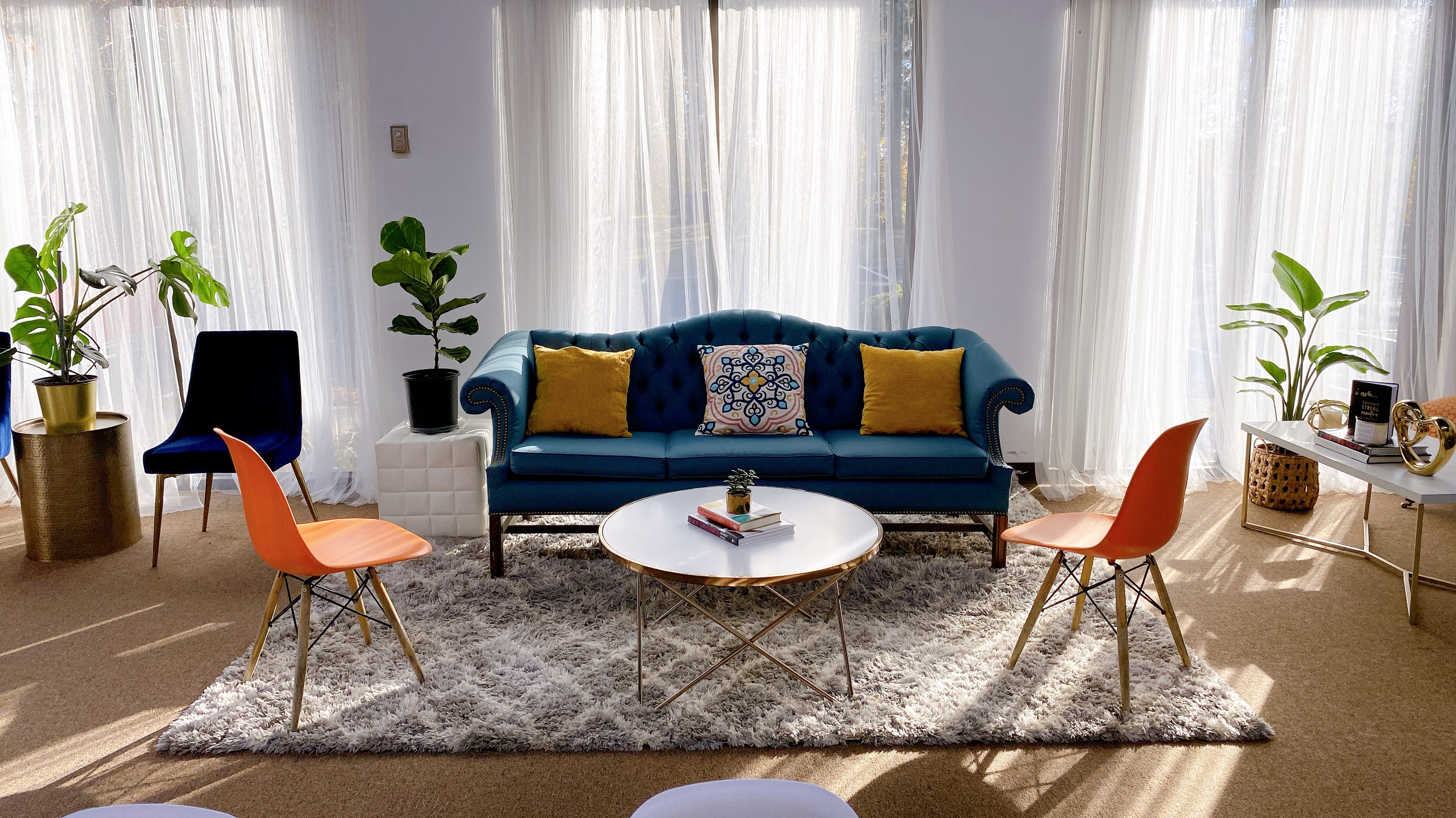 A bright living room features a blue tufted couch with yellow pillows, two orange chairs, a white circular coffee table, and large windows allowing light to filter through sheer curtains.