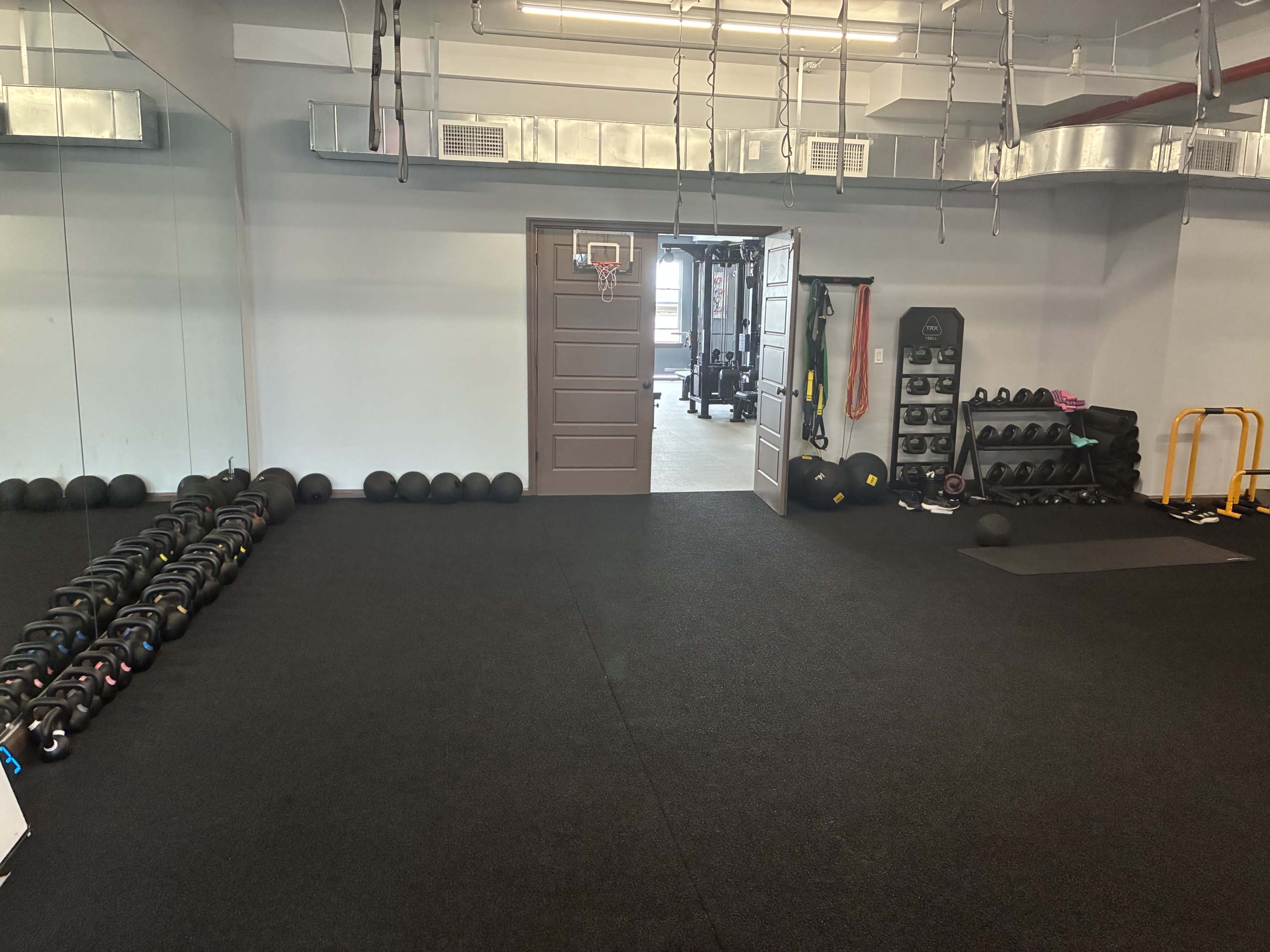 The image shows a fitness room with a variety of weights, a small basketball hoop, and an open door leading to another workout area.