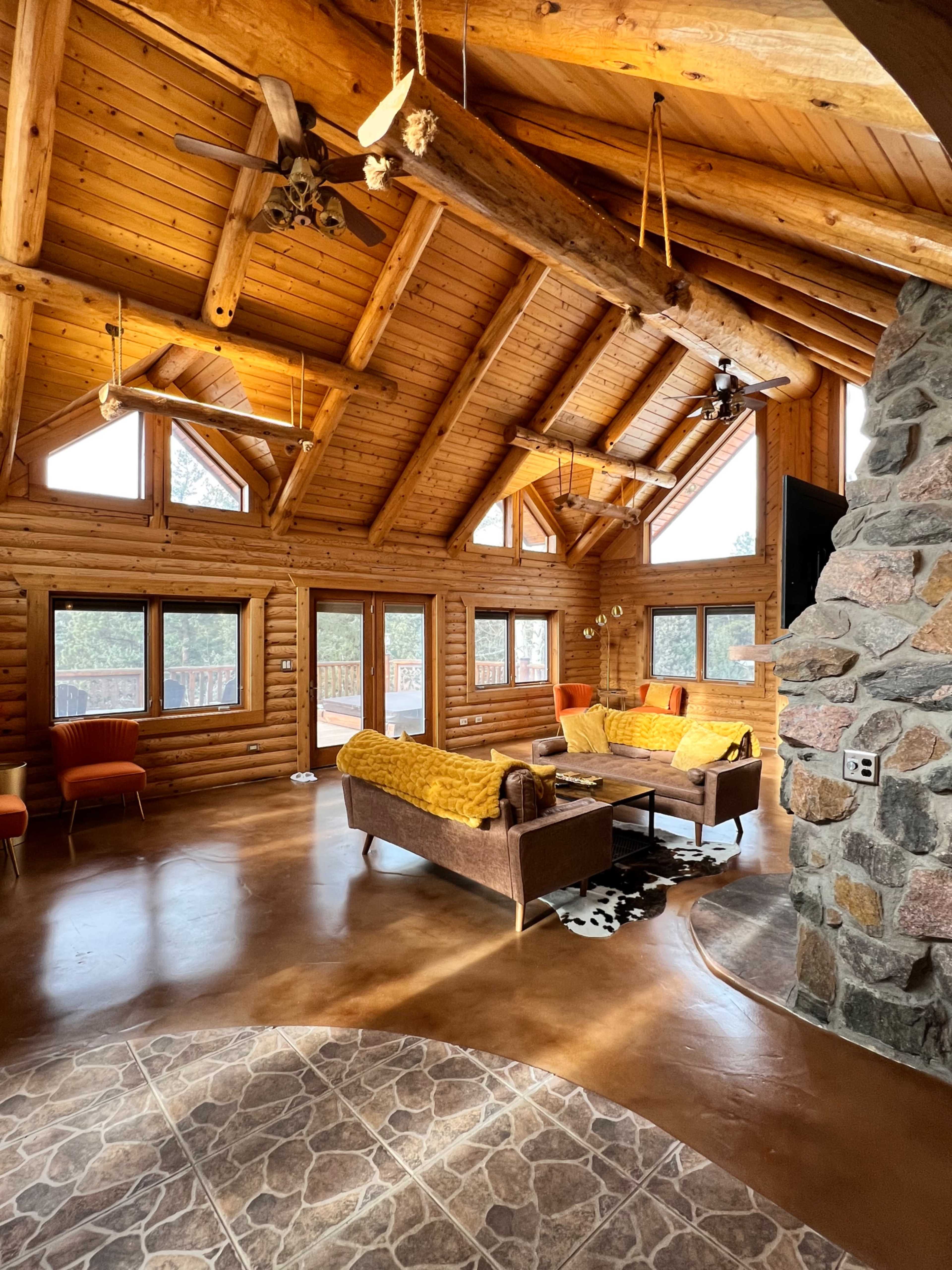 The interior of a log cabin features a high, wooden-beamed ceiling, large windows, and rustic furniture arranged around a stone fireplace.