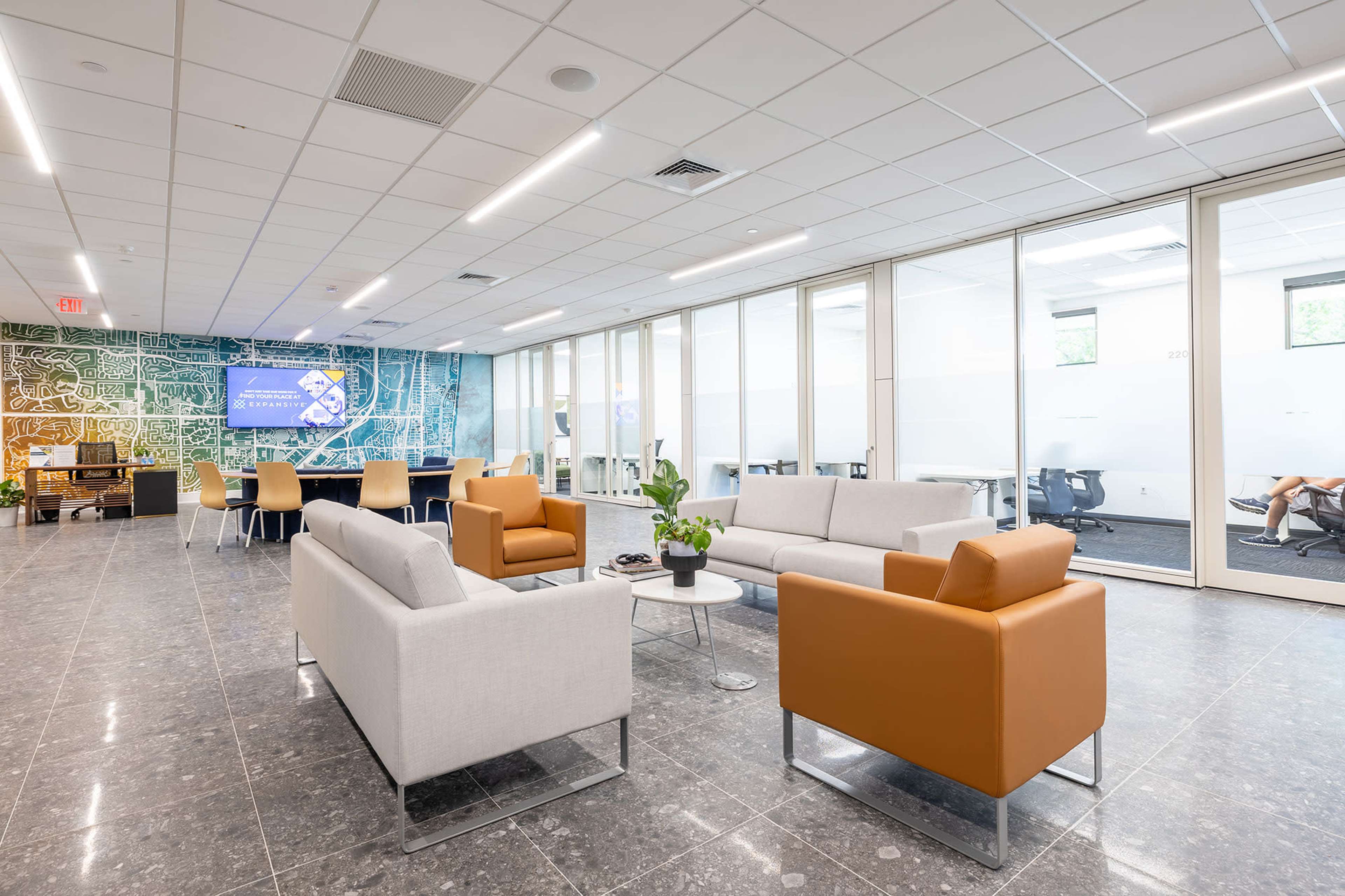 The image shows a modern office lounge area with gray and orange seating, large windows, and a wall featuring a colorful map.