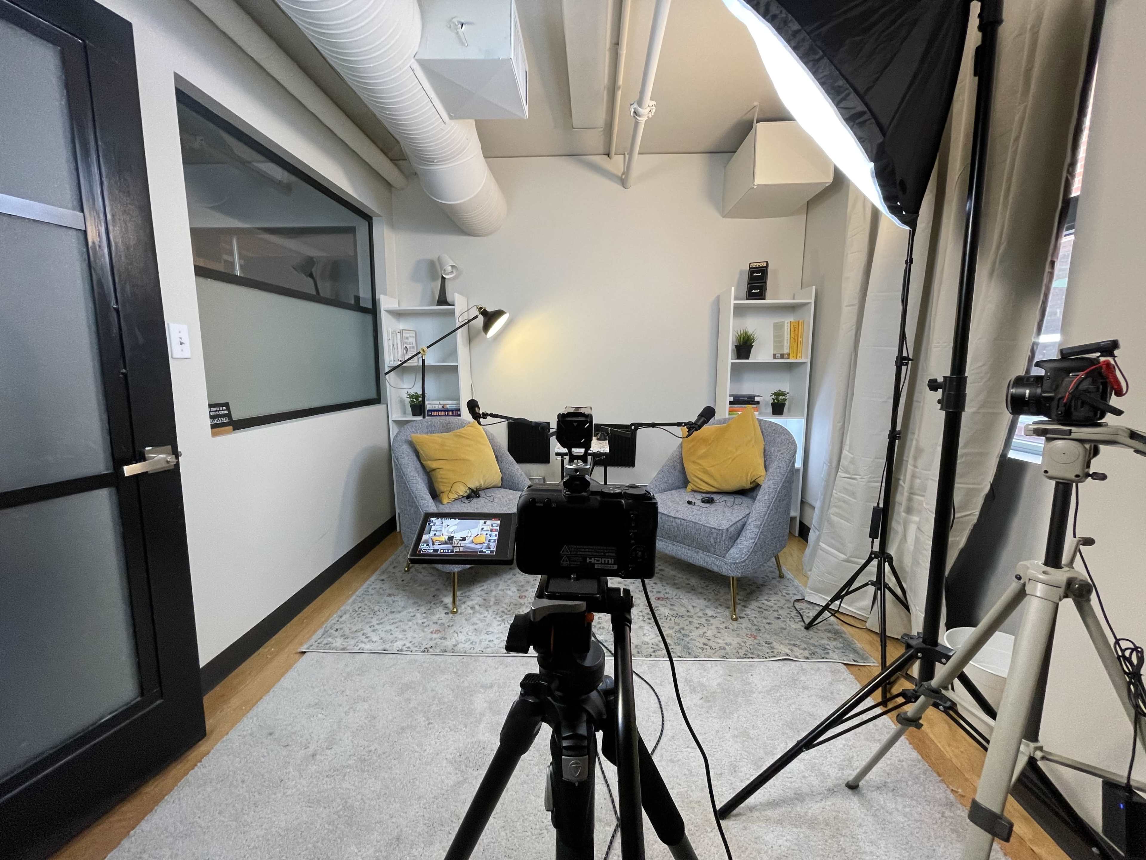 The image shows a well-lit small studio setup featuring a camera on a tripod facing two yellow-cushioned chairs with a small table between them, surrounded by a minimalist decor.