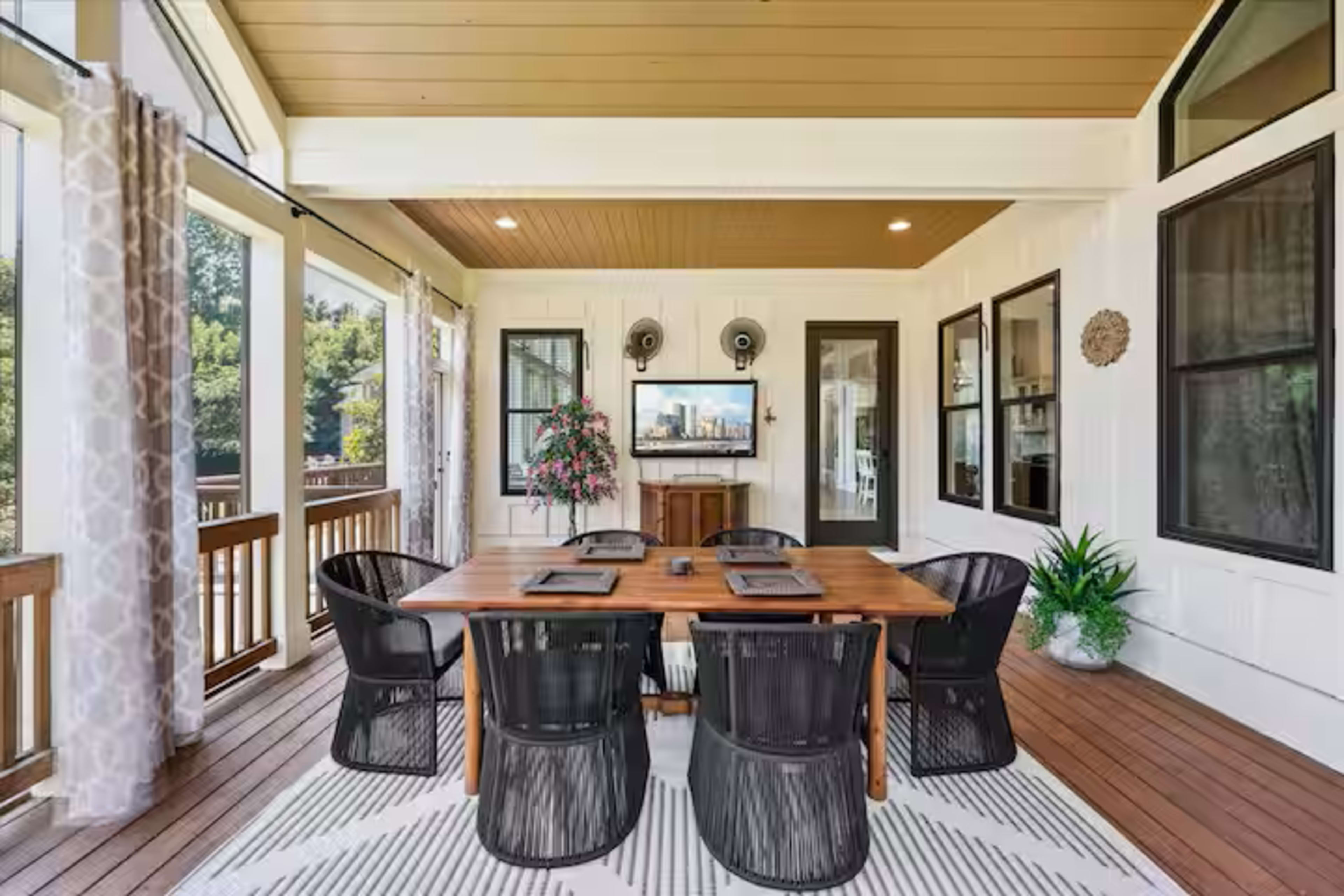 The image shows a covered outdoor dining area with a wooden table, black chairs, and a mounted television on the wall.
