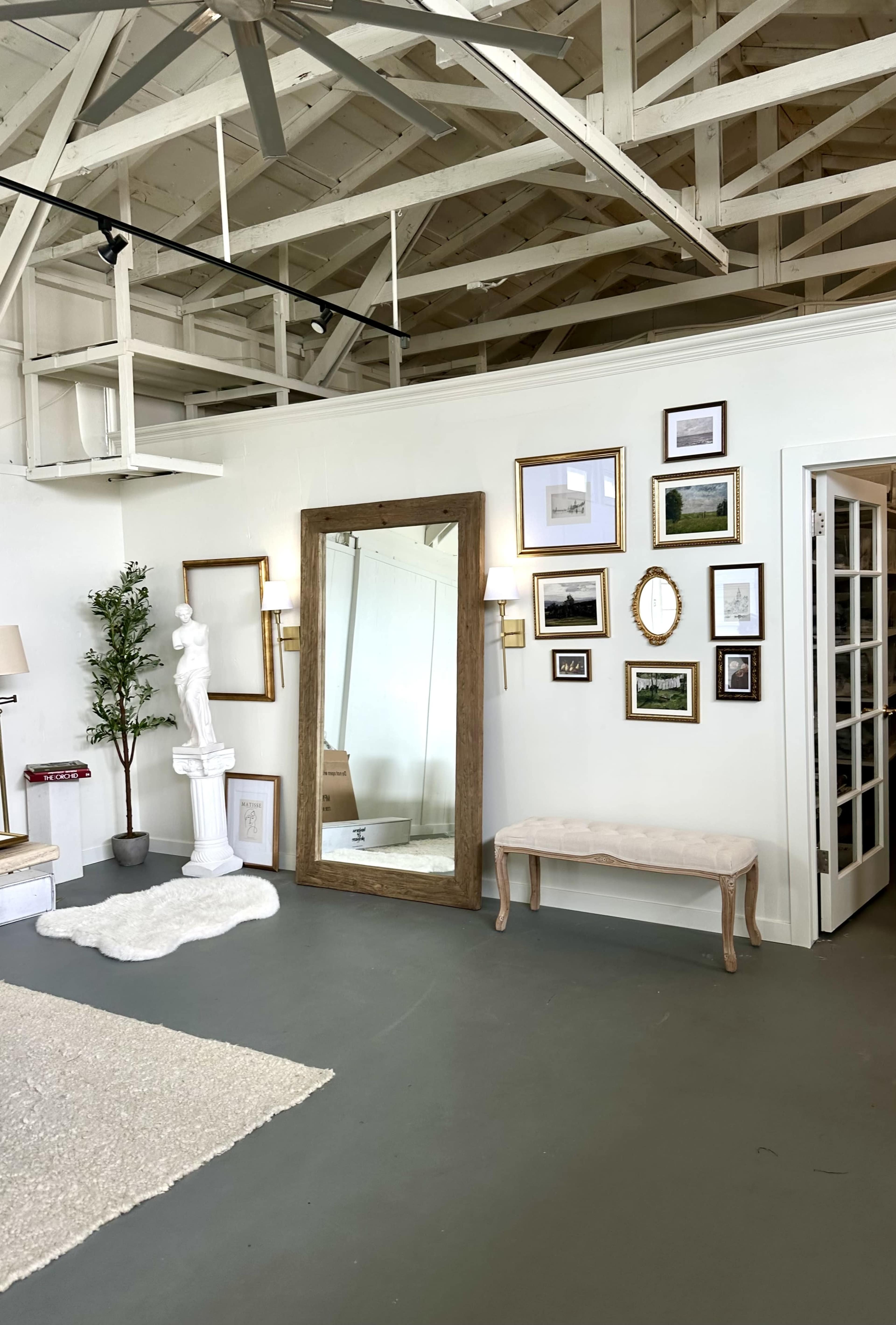 The image shows a spacious room with a large mirror, various framed pictures on the wall, a statue, and a bench in front of a glass door.