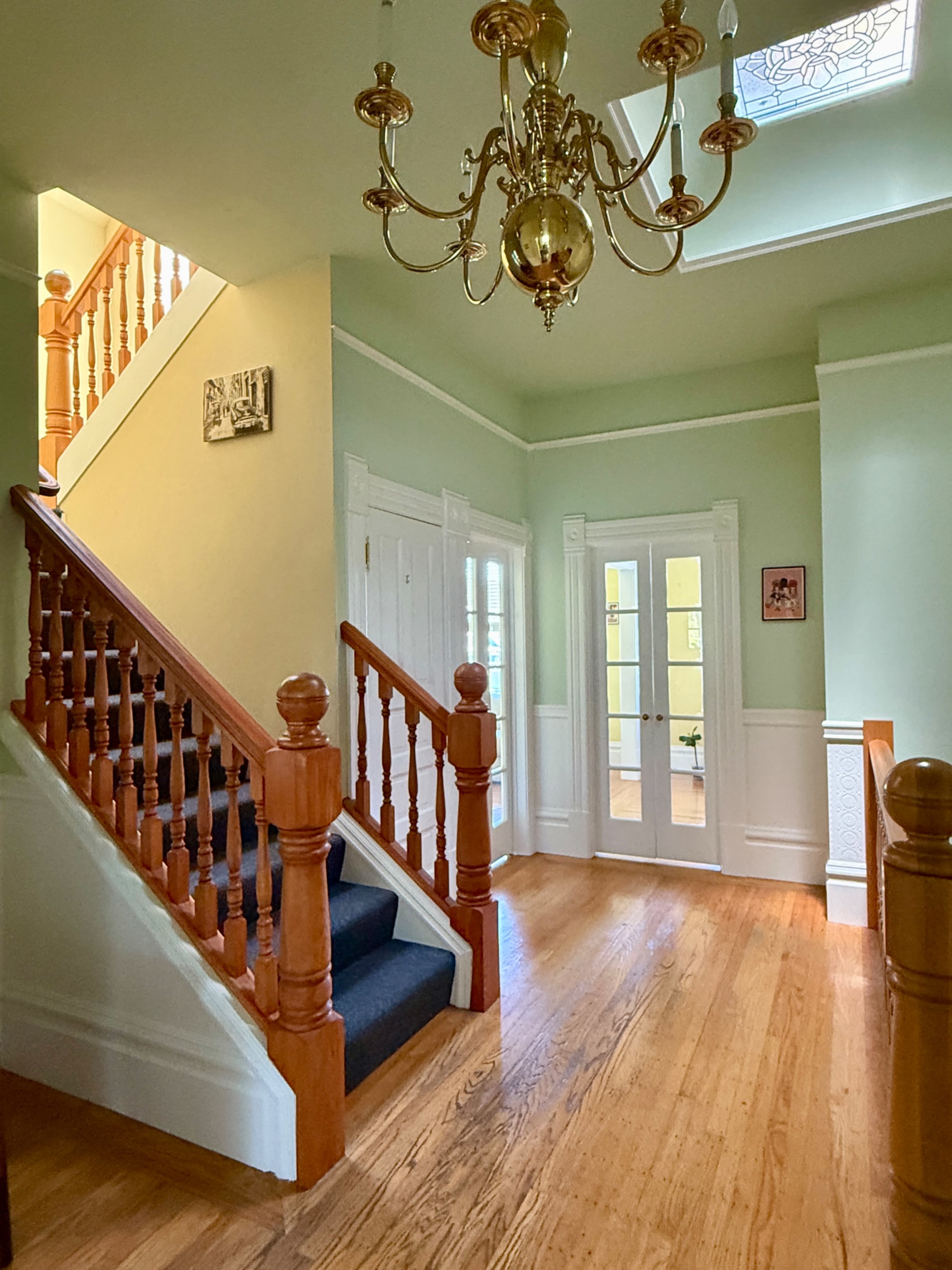 The image shows a well-lit foyer with a wooden staircase, a chandelier, and large double doors leading to another room.