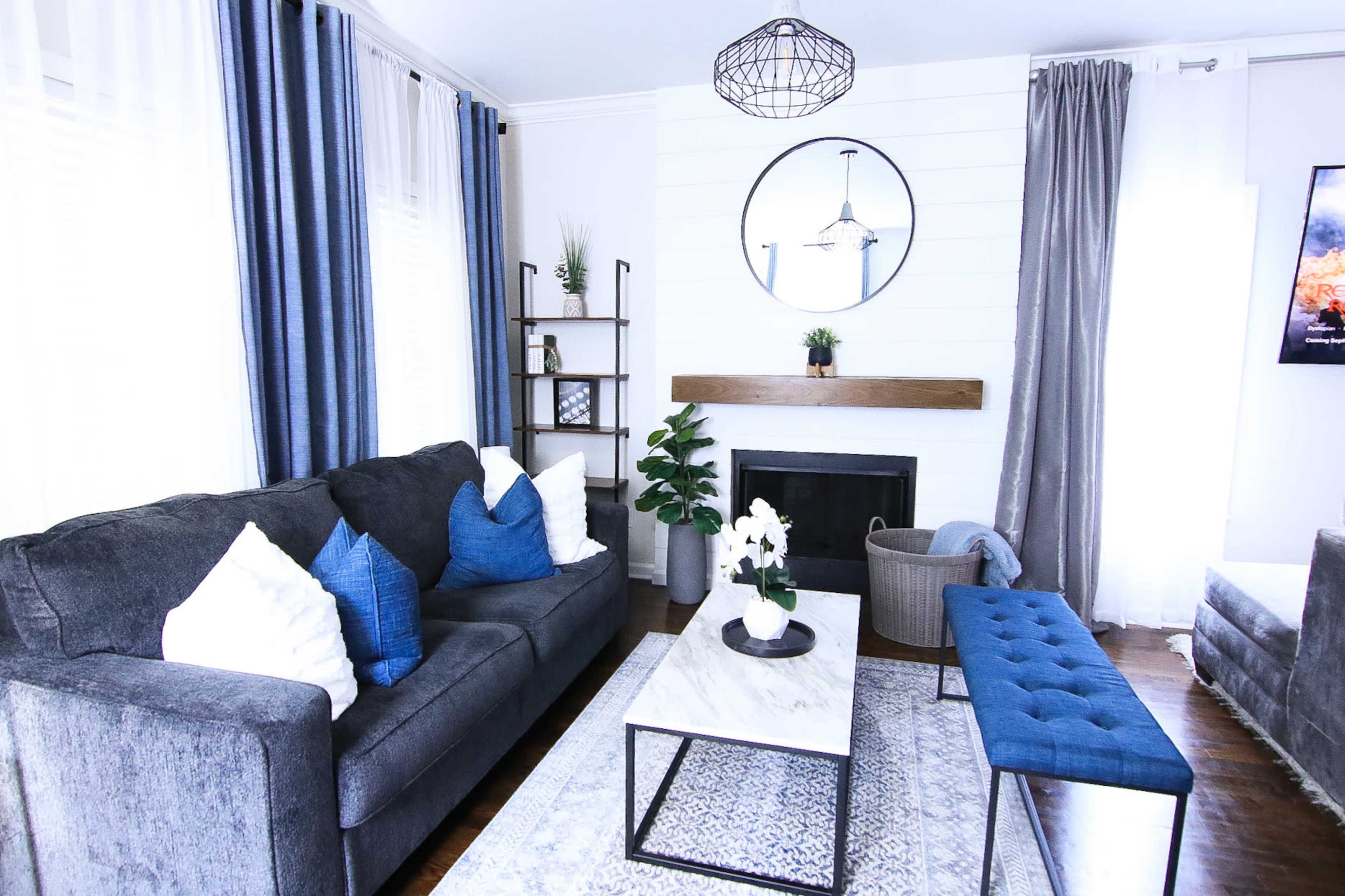 A modern living room features a gray sofa with blue accent pillows, a marble coffee table, and a fireplace surrounded by decorative elements and plants.