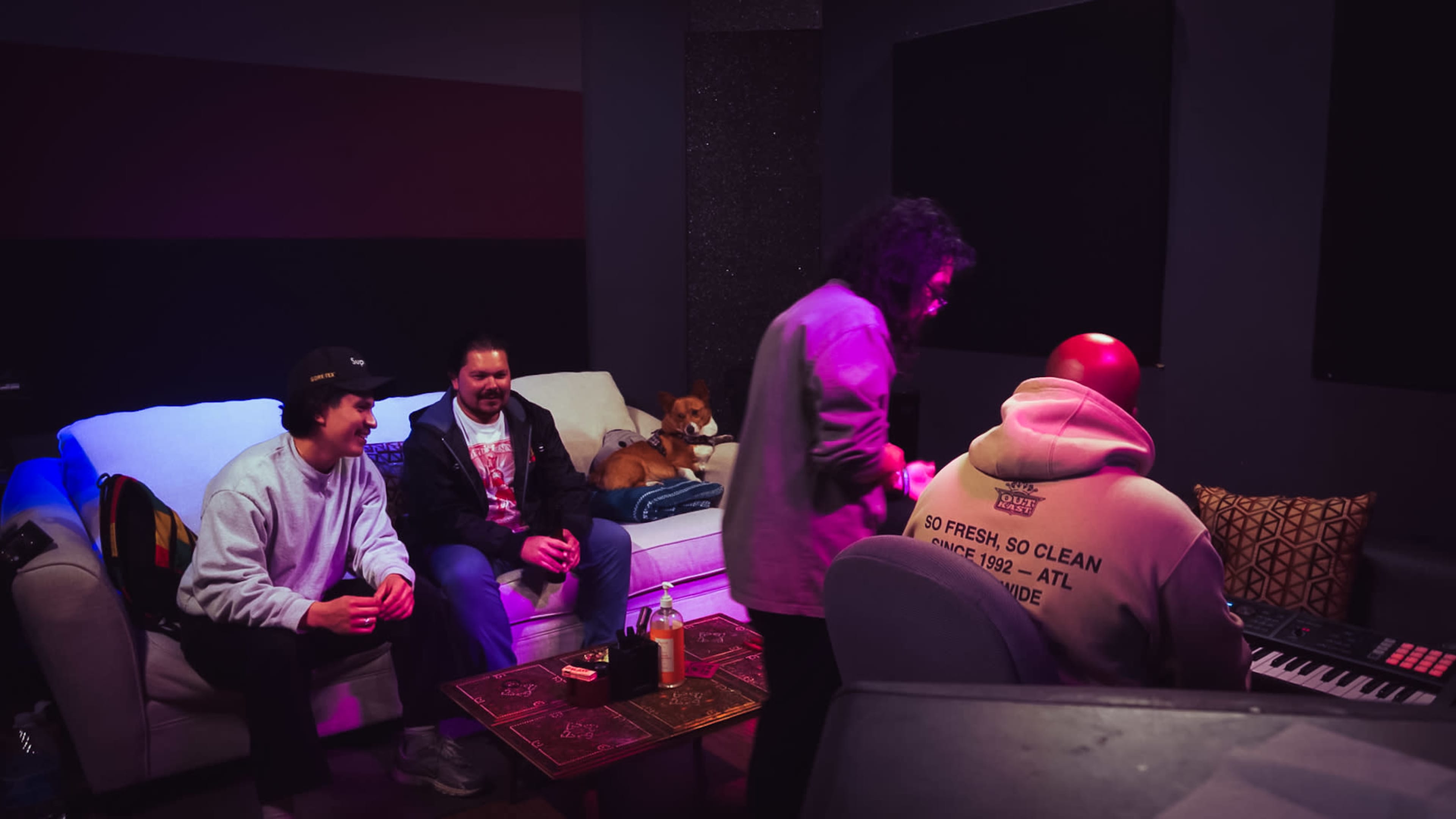 A group of four people is gathered in a music studio, with three seated on a couch and one playing a keyboard, while a dog rests nearby.