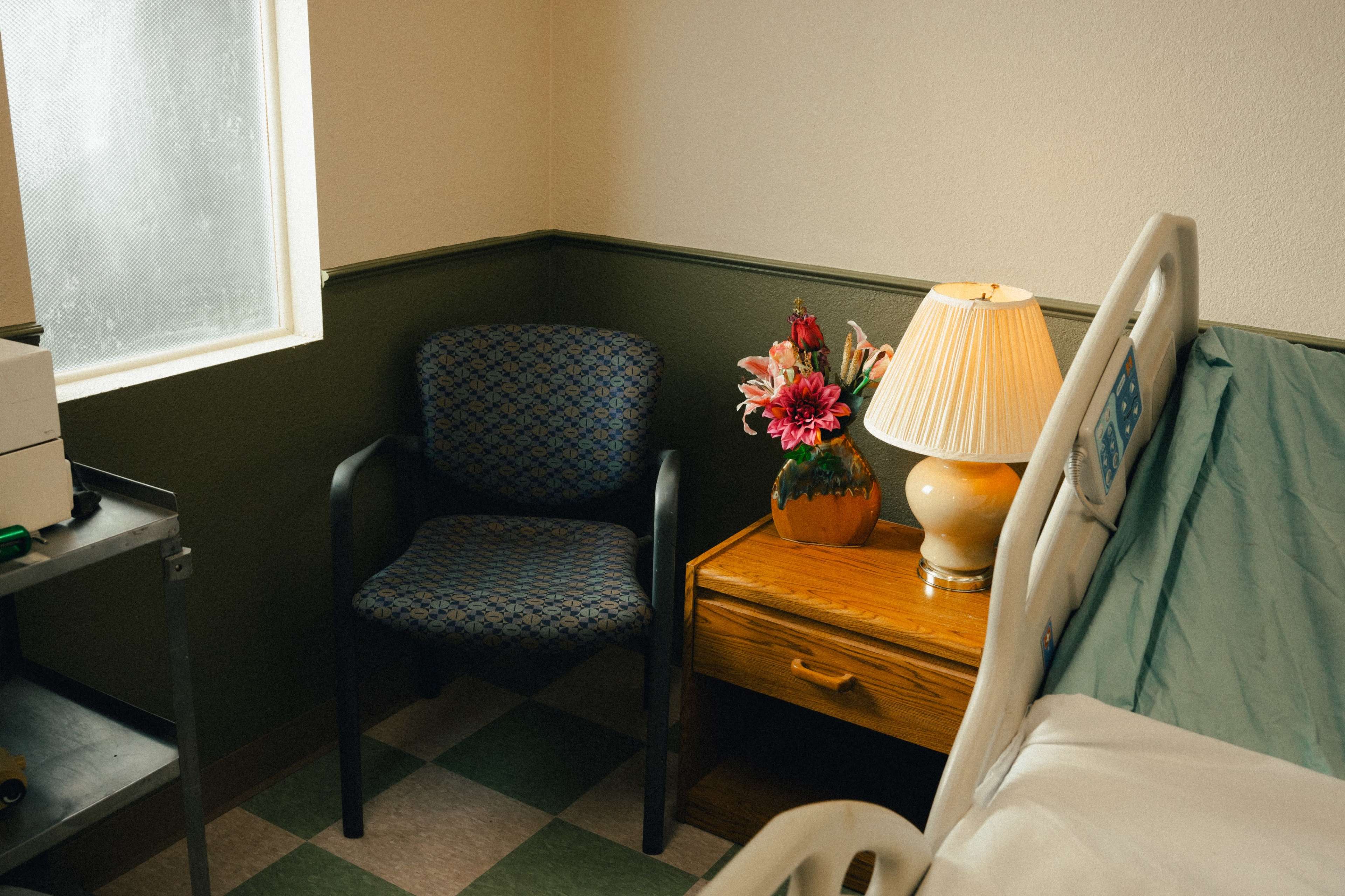 A hospital room features a bed, a nightstand with a lamp and flowers, a chair, and a small table near a window.