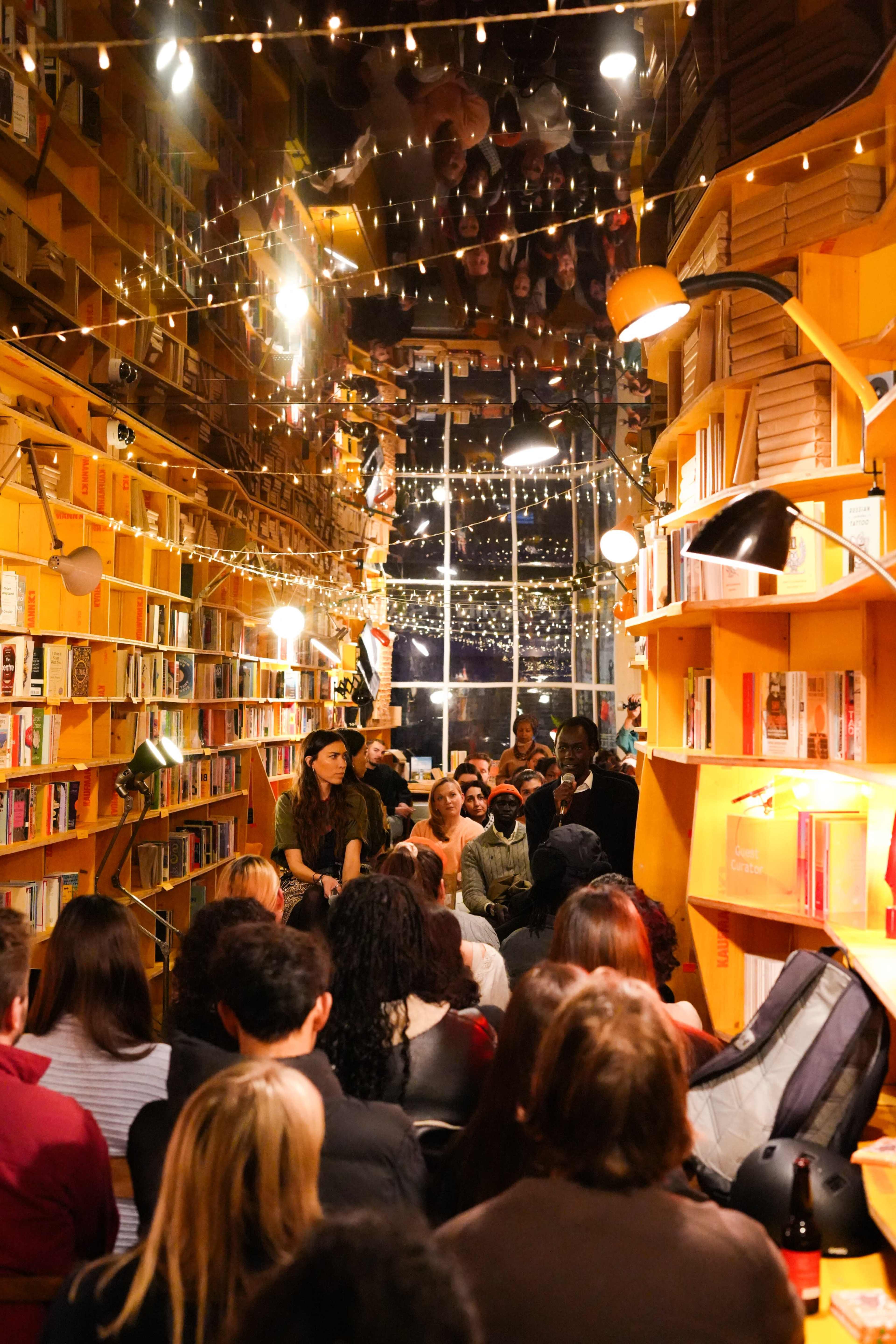 The image shows a crowded bookshop with bookshelves on either side, where an audience is attending an event under string lights reflecting in the windows.