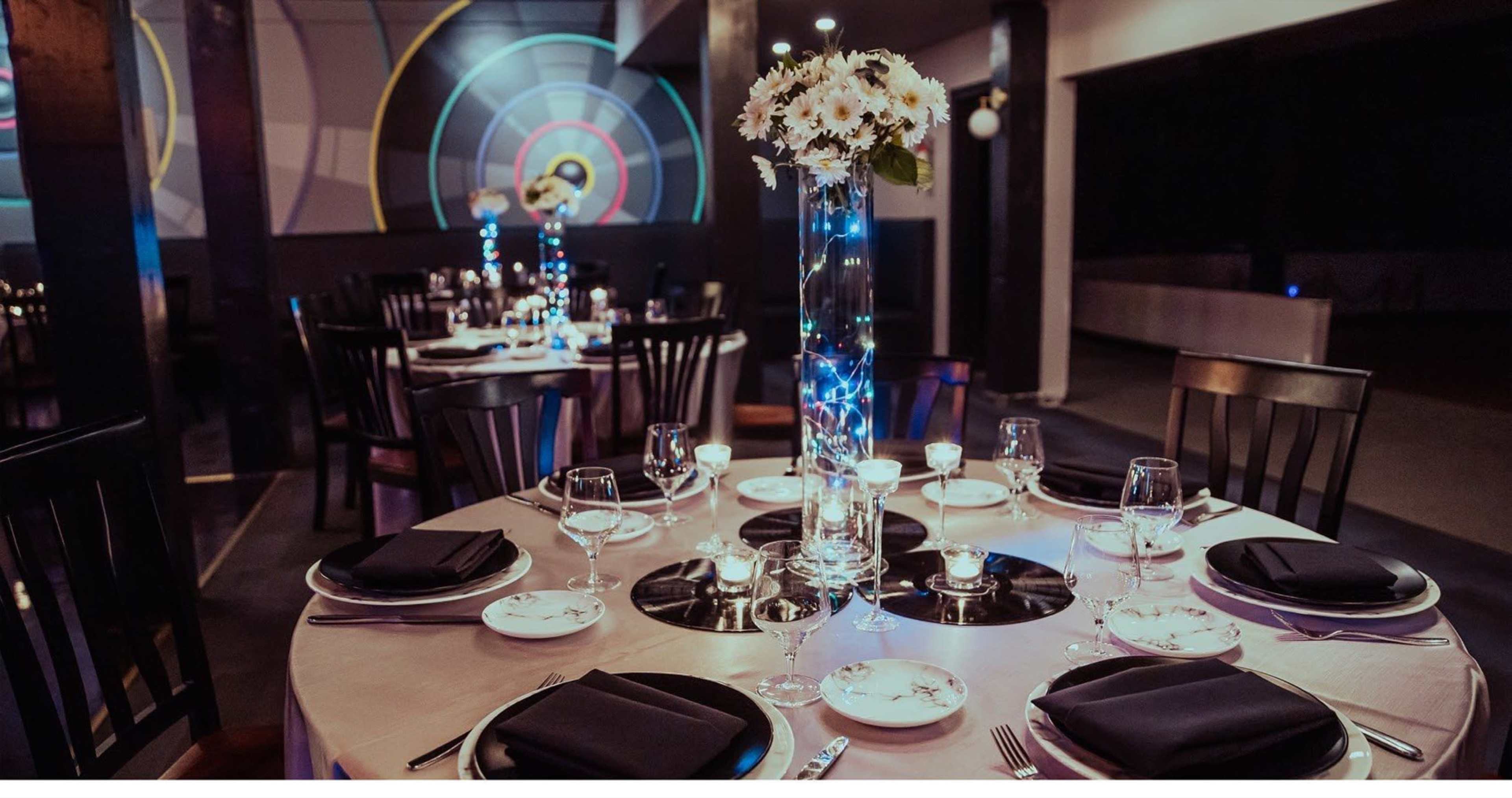 A round dining table is set elegantly with black napkins, wine glasses, and a tall floral centerpiece, surrounded by dark wooden chairs in a modern restaurant setting.