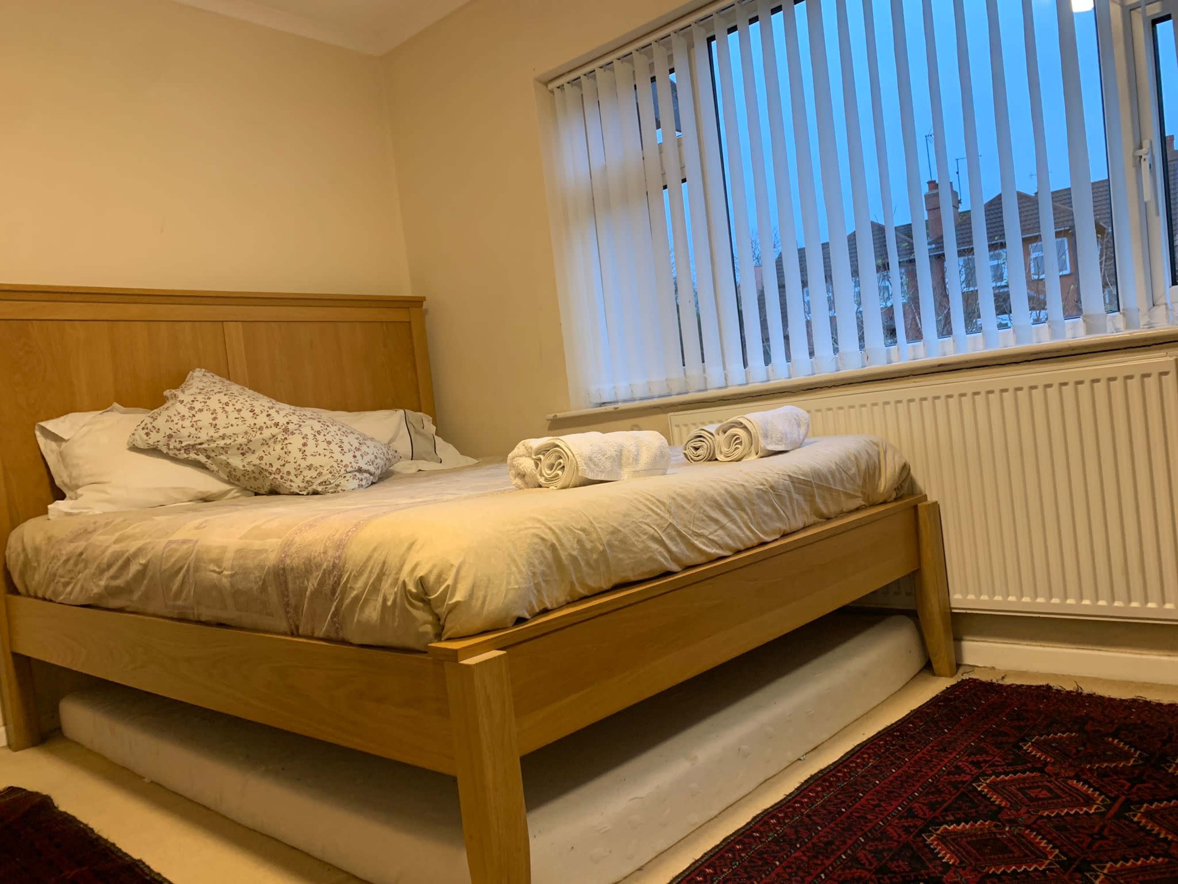The image shows a simple bedroom with a wooden bed, three rolled towels on the bed, and vertical blinds partially drawn over a window.