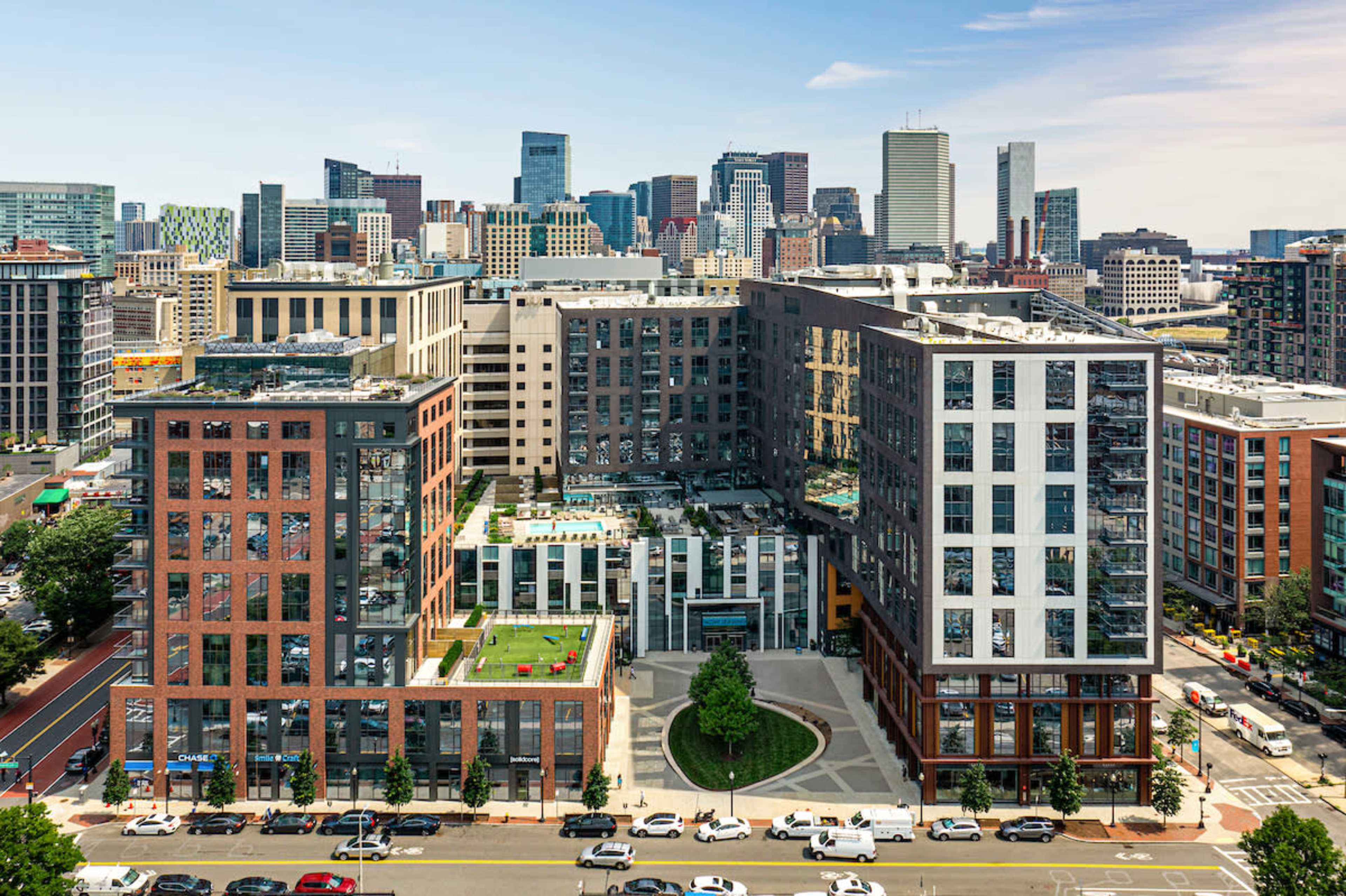 A modern urban residential complex with several multi-story buildings and green spaces, set against a backdrop of a city skyline with high-rise structures.