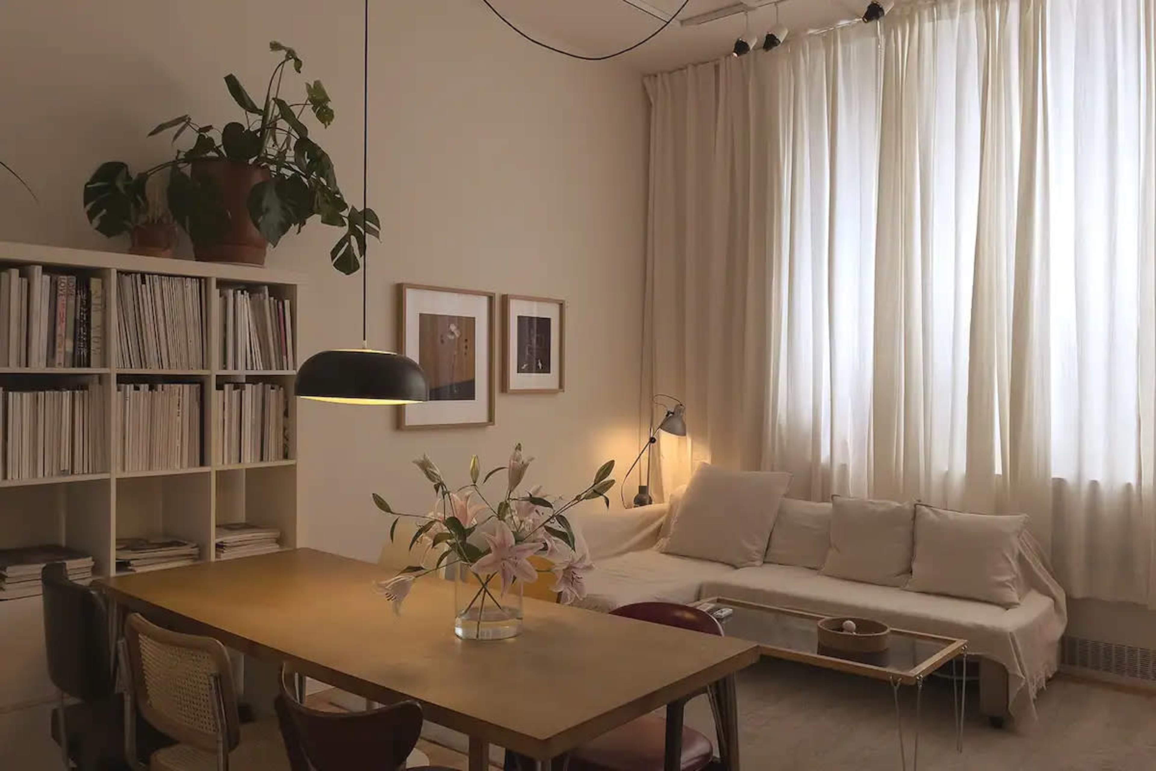 A cozy living space features a dining table with a flower arrangement, a light-colored sofa, large curtains, and a bookshelf filled with magazines.