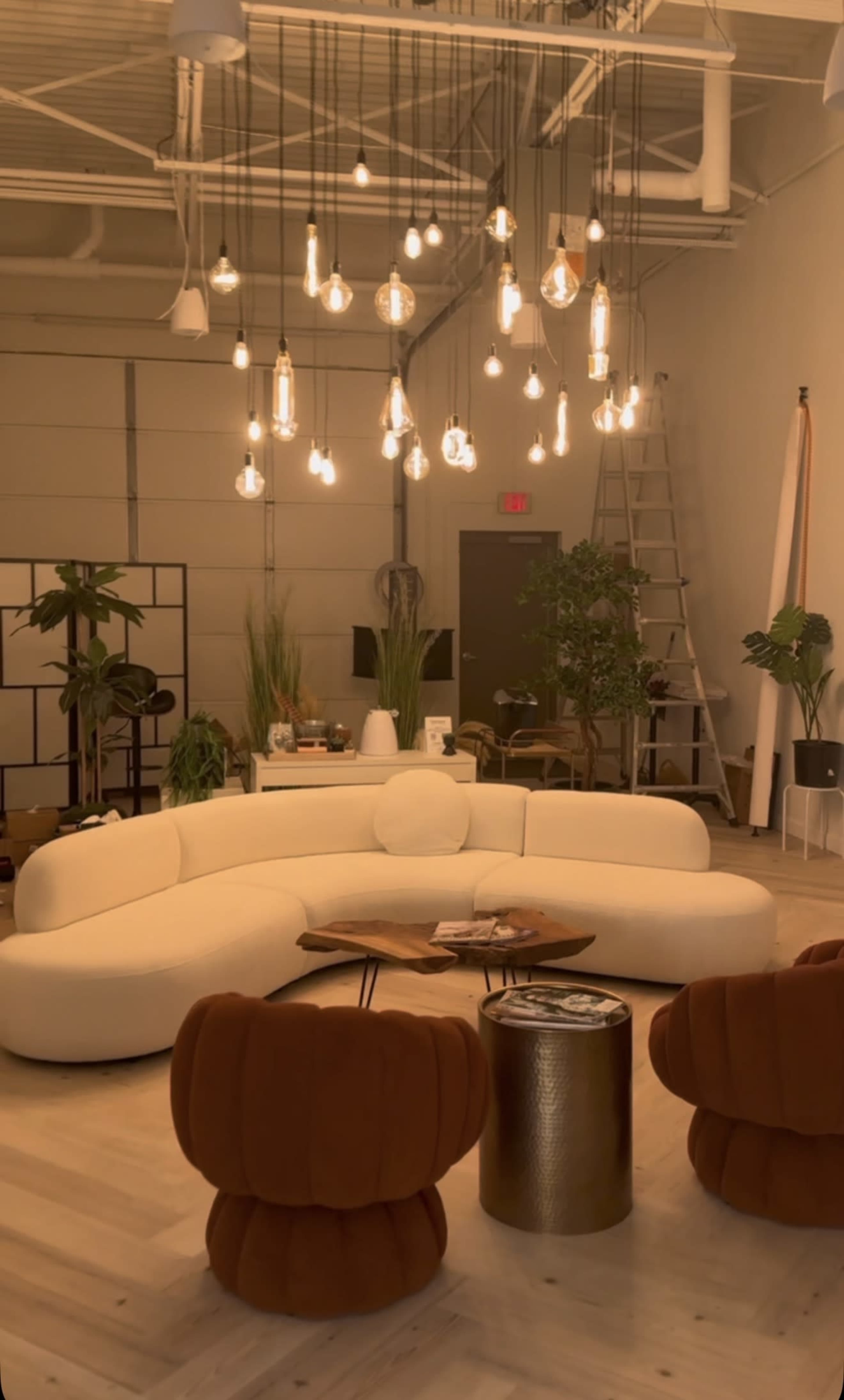 The image shows a modern room featuring a white curved sofa, two brown upholstered chairs, a round coffee table, and various plants, all illuminated by numerous hanging light bulbs.