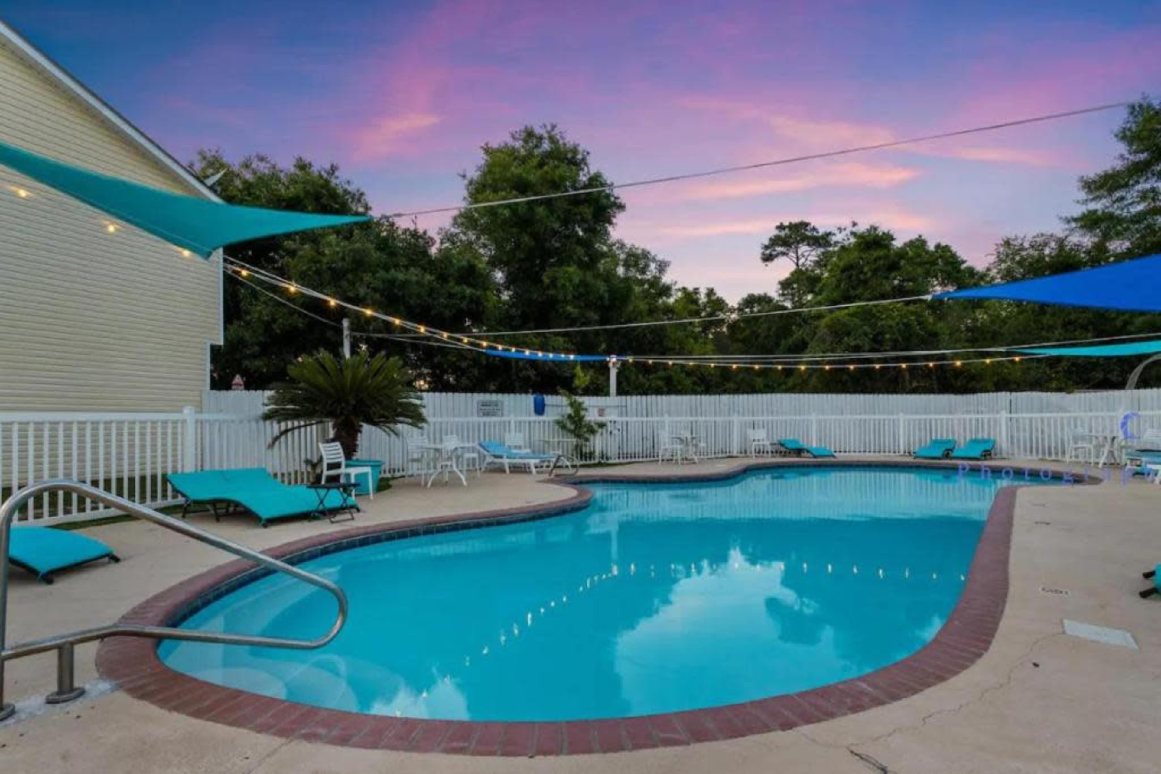 A swimming pool surrounded by lounge chairs and shaded by blue canopies, with trees and a white fence in the background.