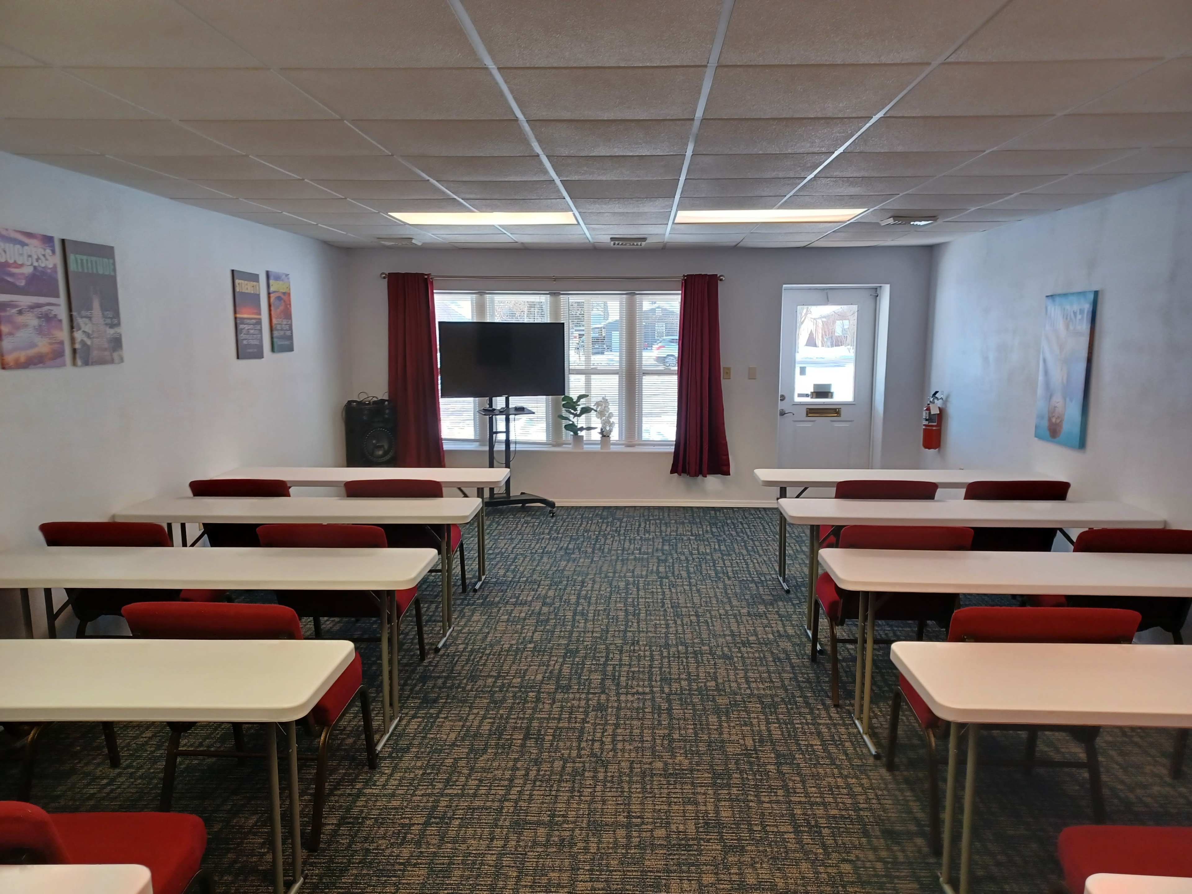 A classroom is set up with rows of tables and chairs facing a large window, featuring a television screen on a stand.