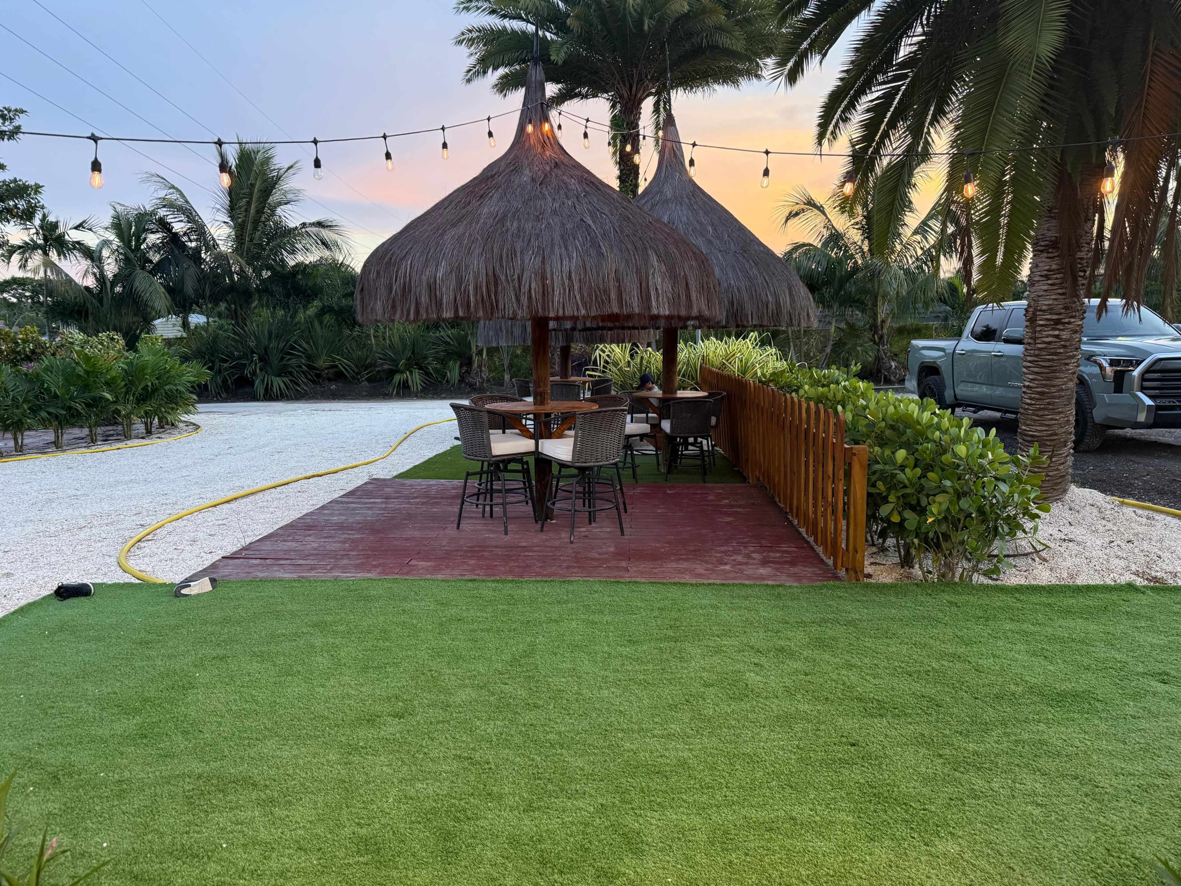 The image shows a thatched-roof gazebo with dining tables surrounded by palm trees and string lights, set against a sunset sky.