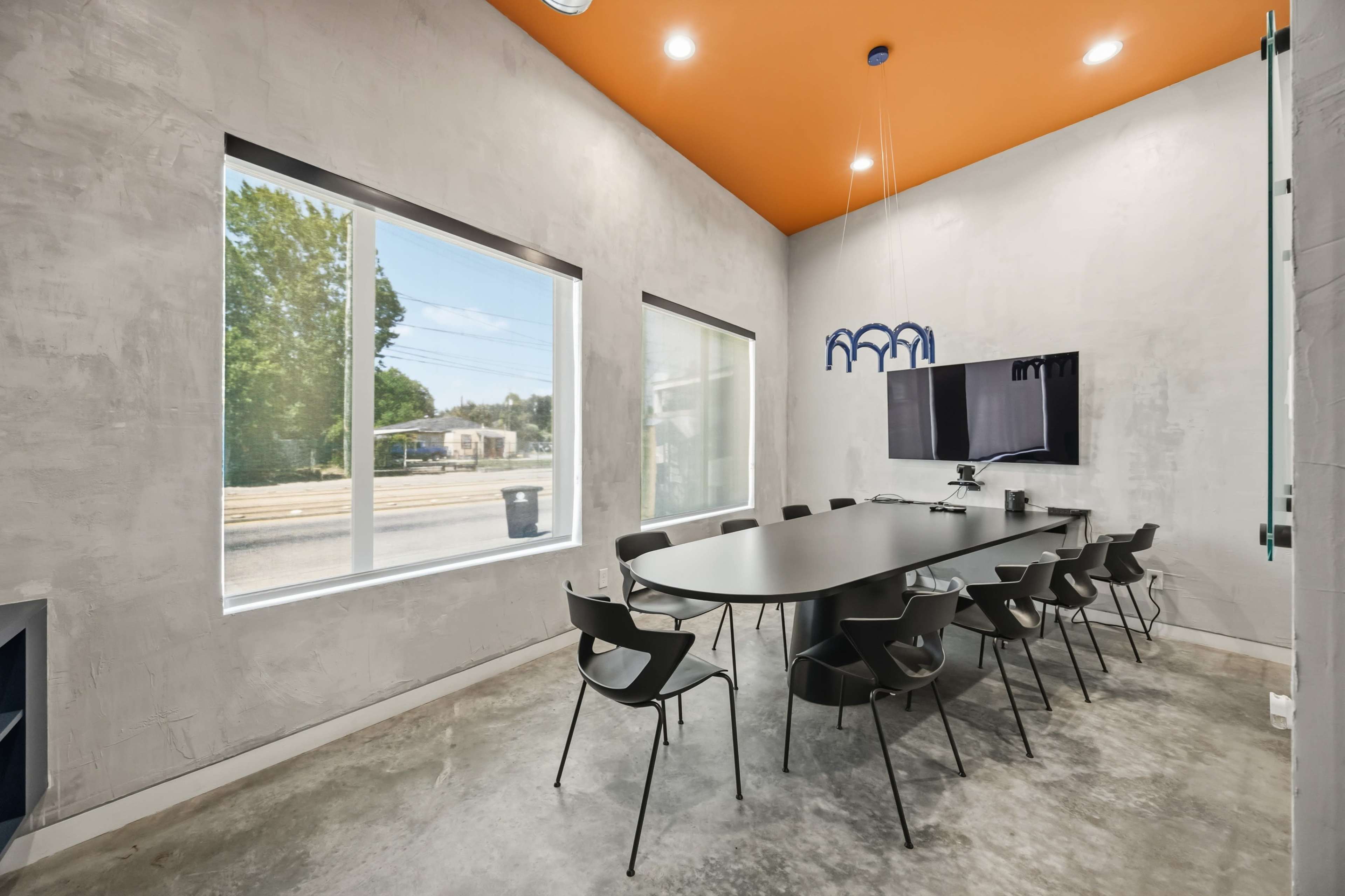 A modern conference room features a long oval table surrounded by black chairs, with large windows letting in natural light and a television mounted on the wall.
