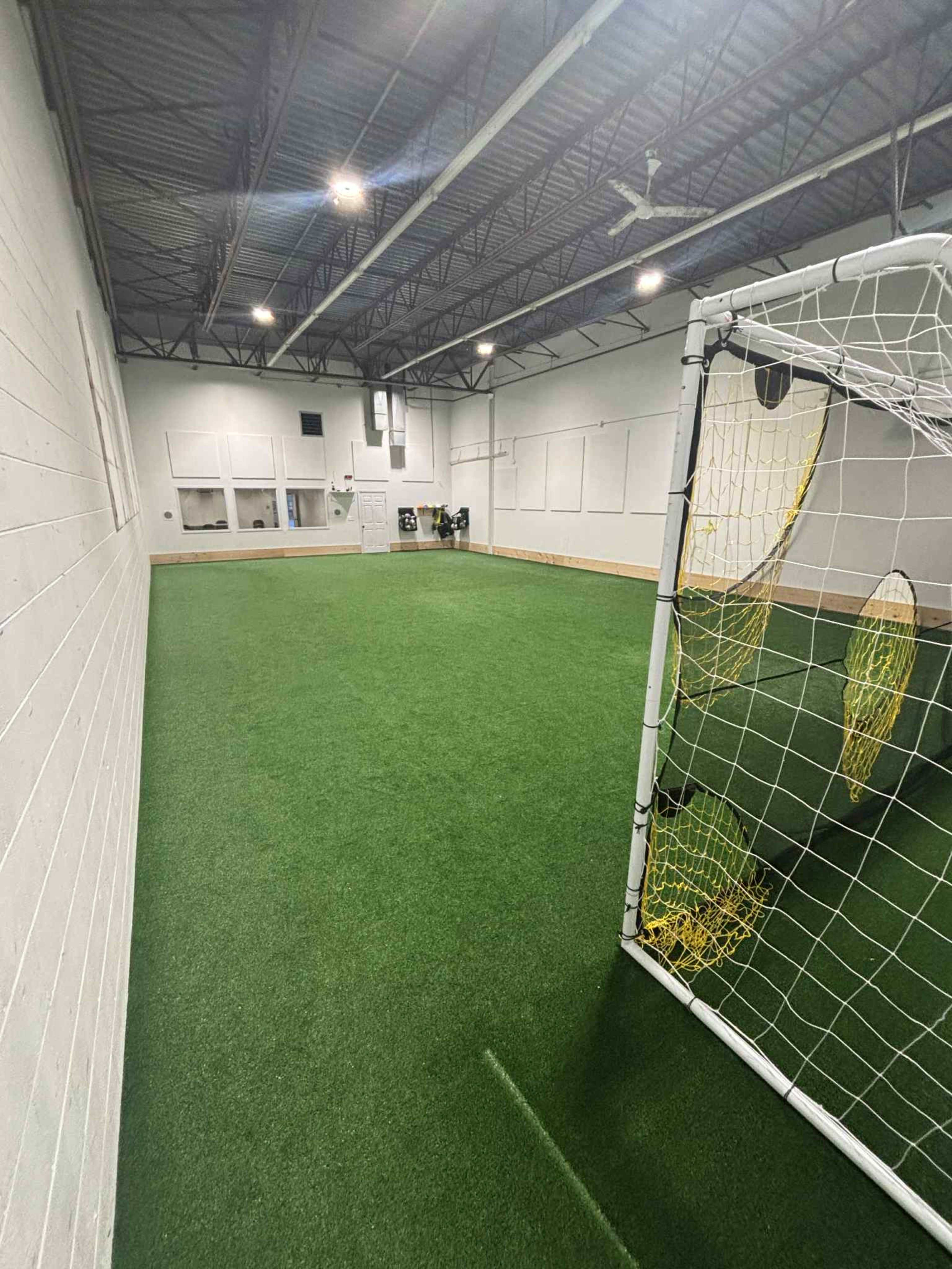 The image shows an indoor soccer training facility featuring artificial turf and a goalpost in the foreground.