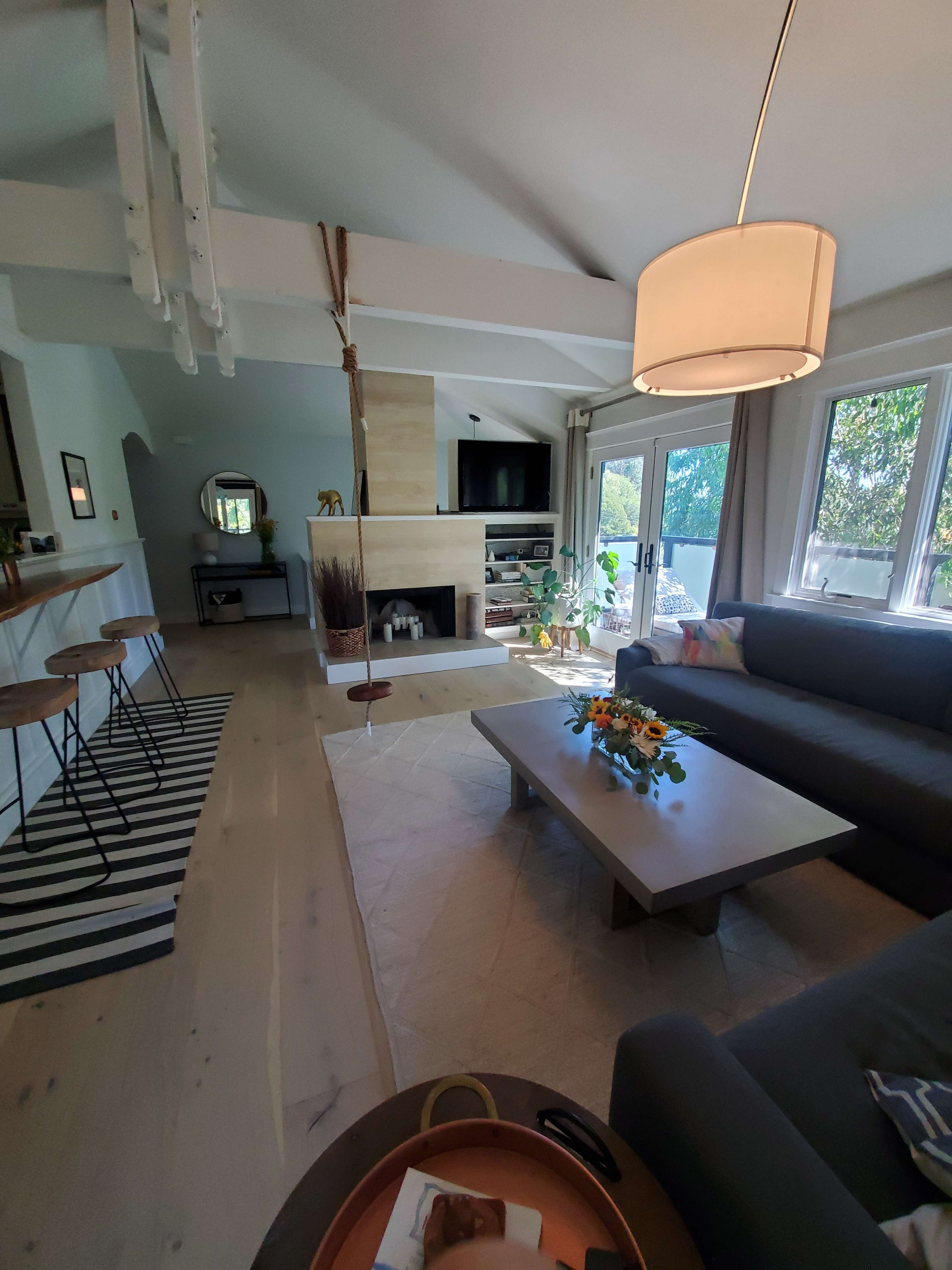 The image shows a modern living room with a sectional sofa, a coffee table, and a fireplace, connected to a kitchen with bar stools and large windows allowing natural light.