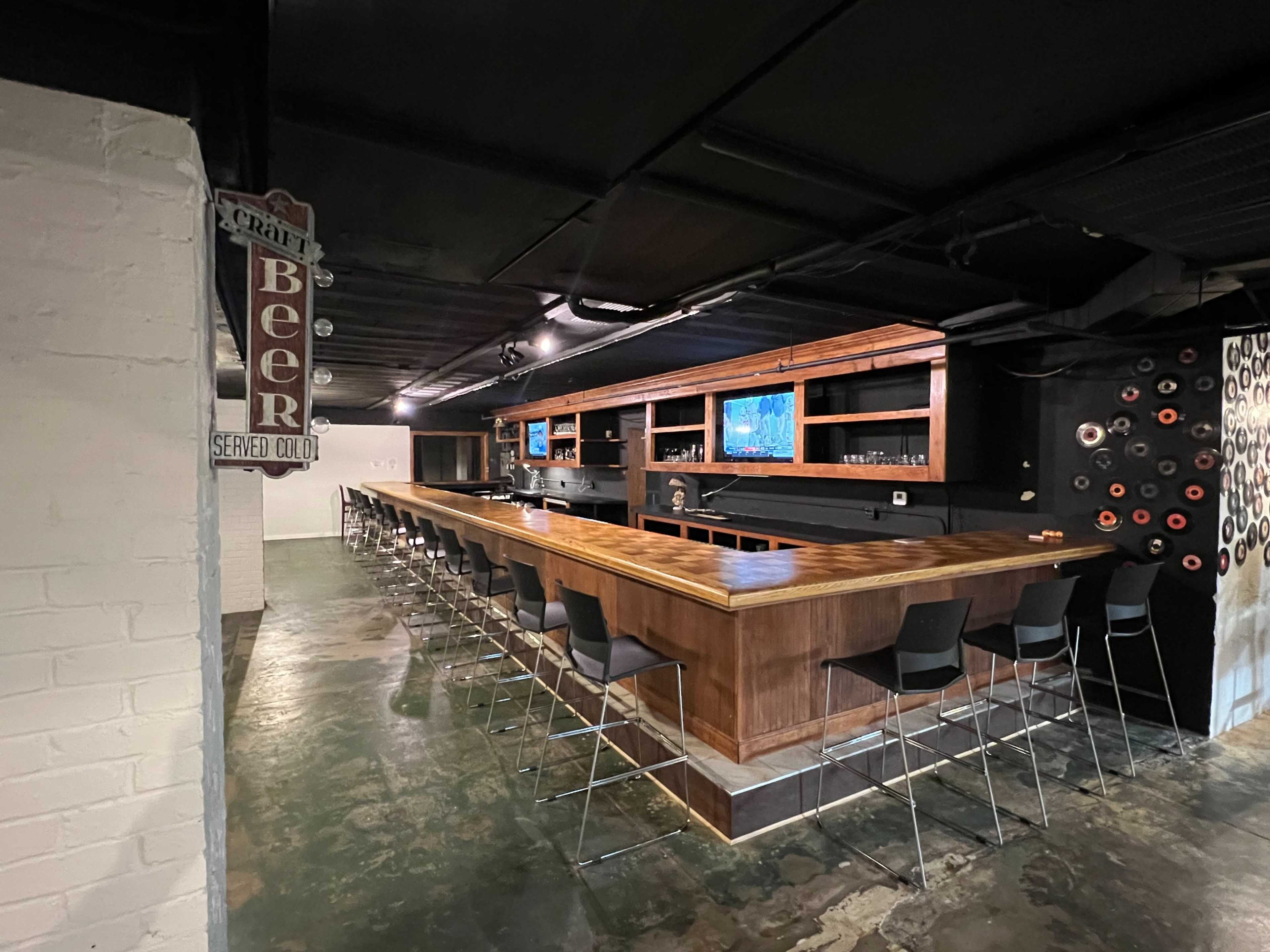 A modern bar with a long wooden counter and high black stools, set in an industrial-style space with exposed walls and dim lighting.