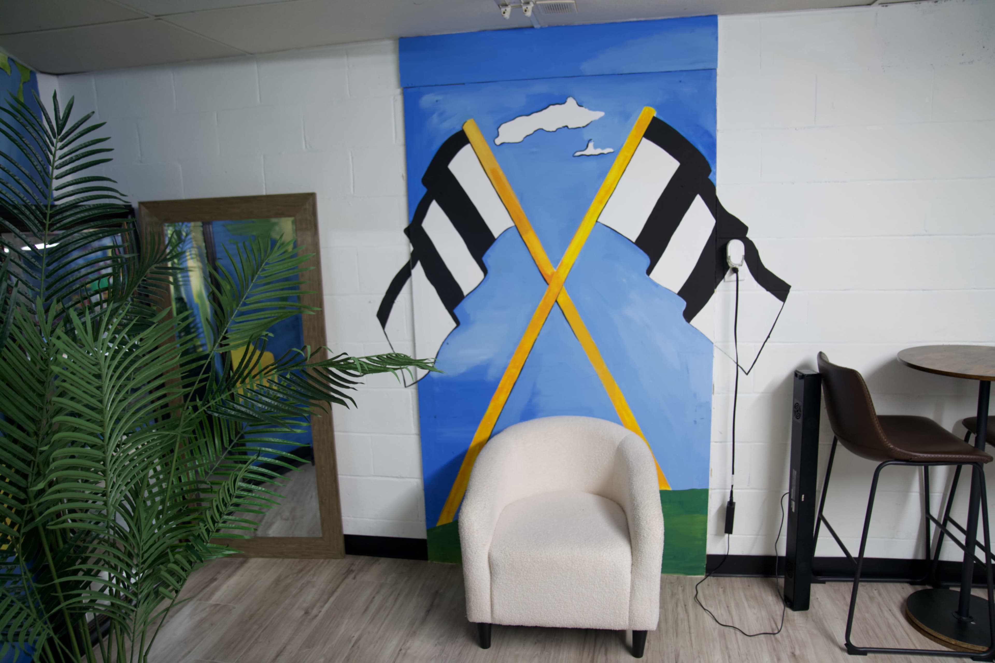 The image shows a minimalist interior space featuring a white chair in front of a mural depicting crossed black and white flags against a blue sky.