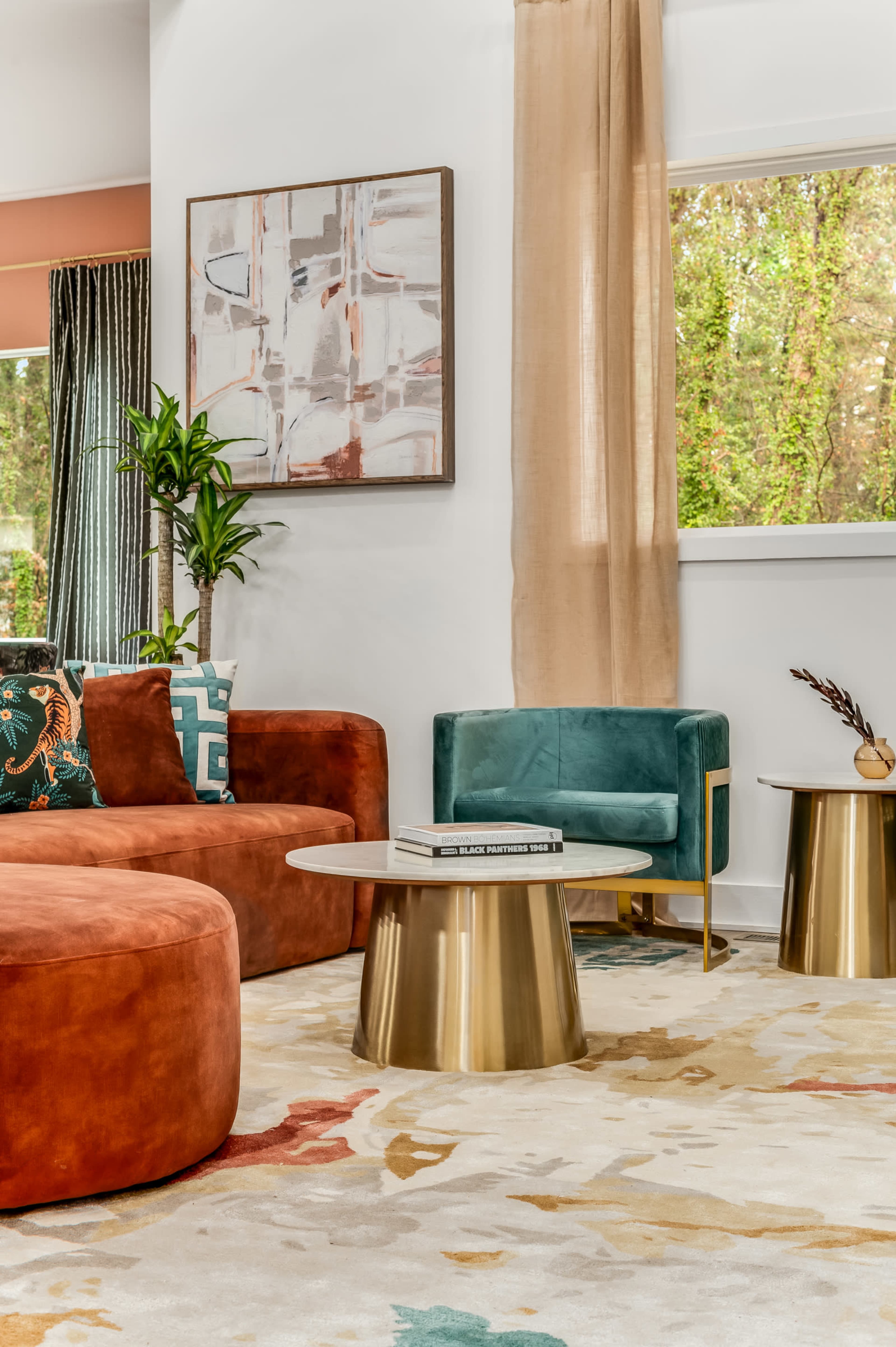 The image shows a modern living room featuring a large rust-colored circular sofa, a teal armchair, a round coffee table with a gold base, and a vibrant area rug.