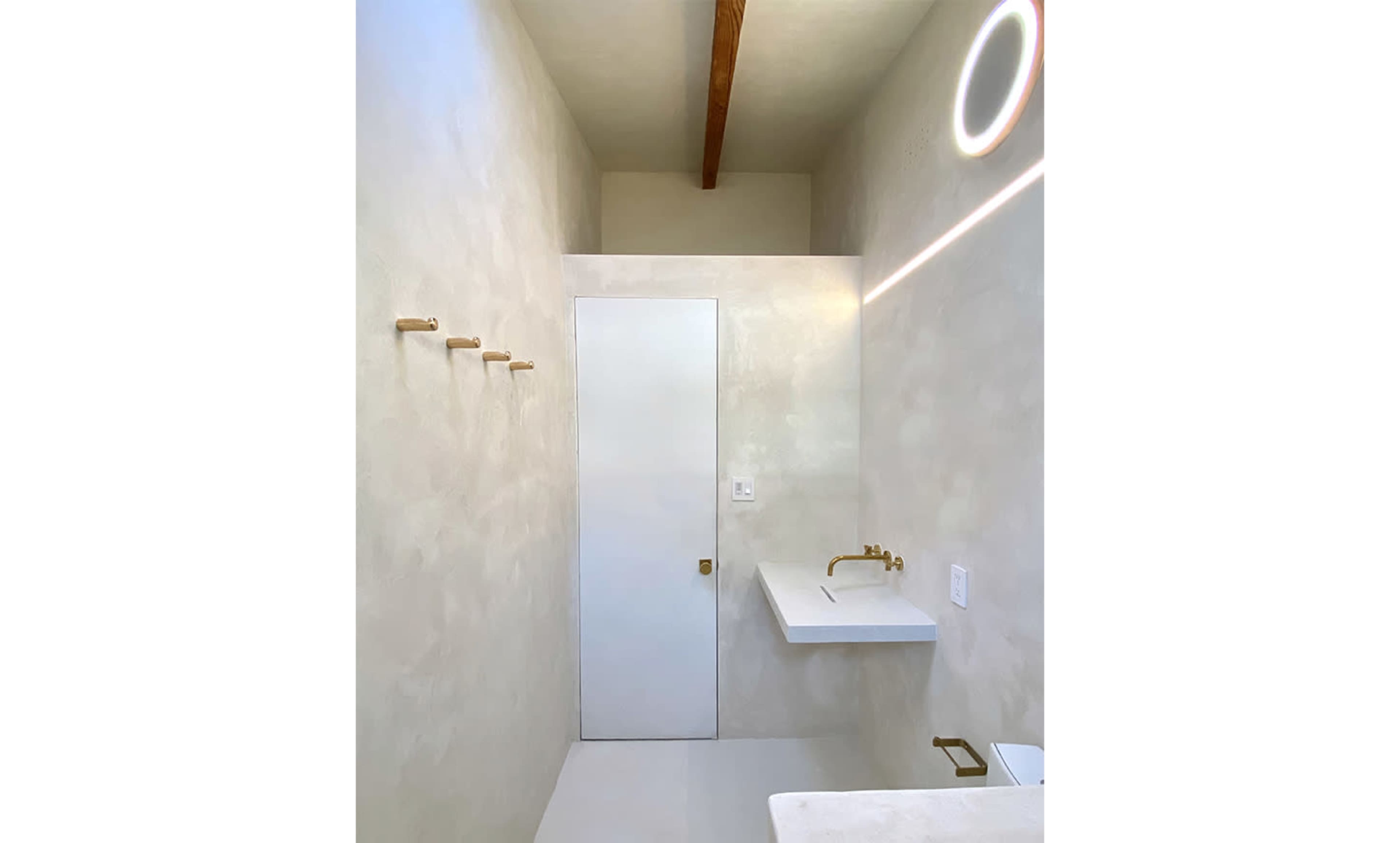 The image shows a minimalist bathroom with a white wall, a wooden beam ceiling, a door, a sink with a gold faucet, and several wooden hooks mounted on the wall.