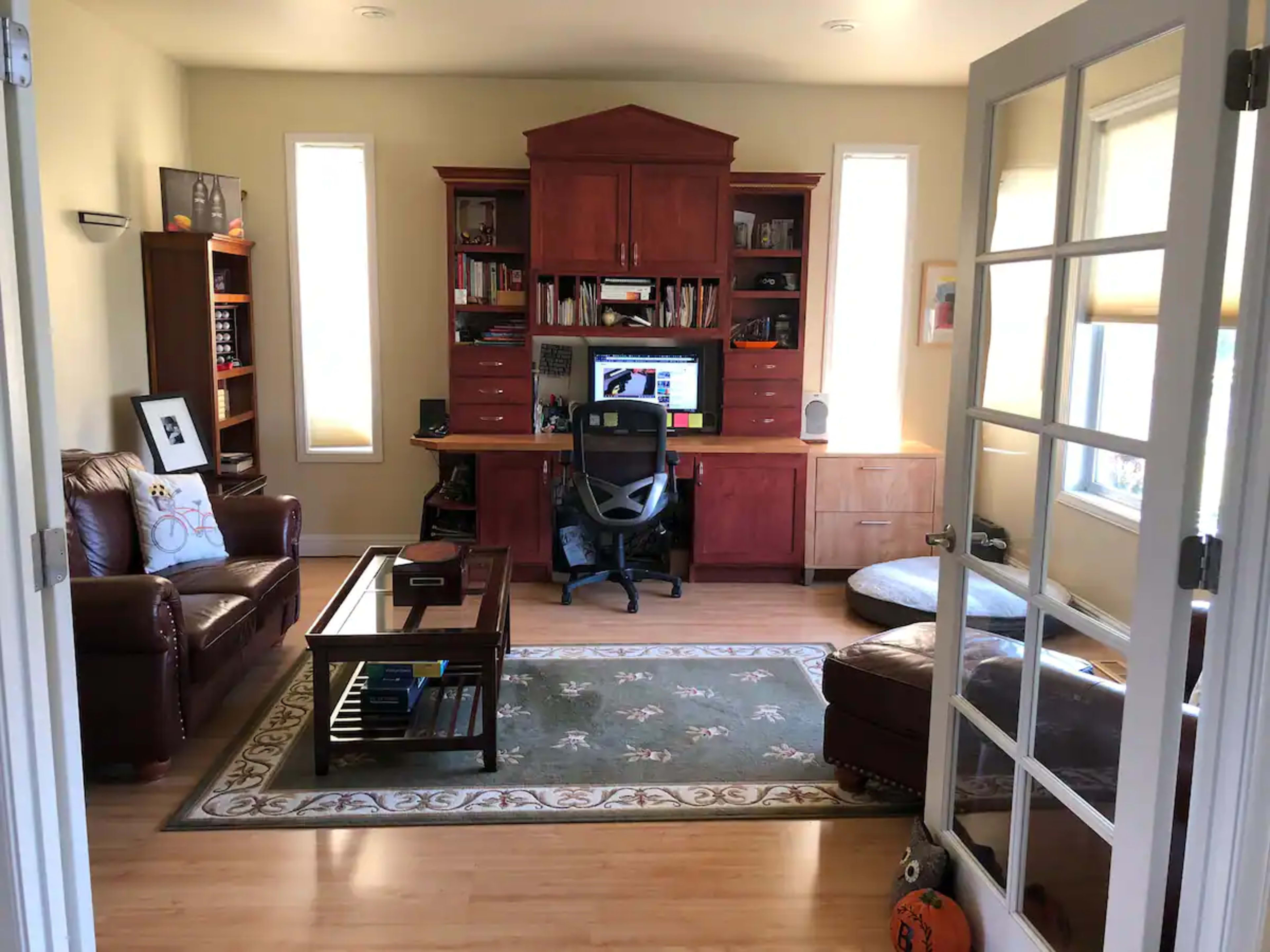 A home office features a wooden desk with a computer, bookshelves, two leather chairs, and a coffee table, all arranged around a rug.