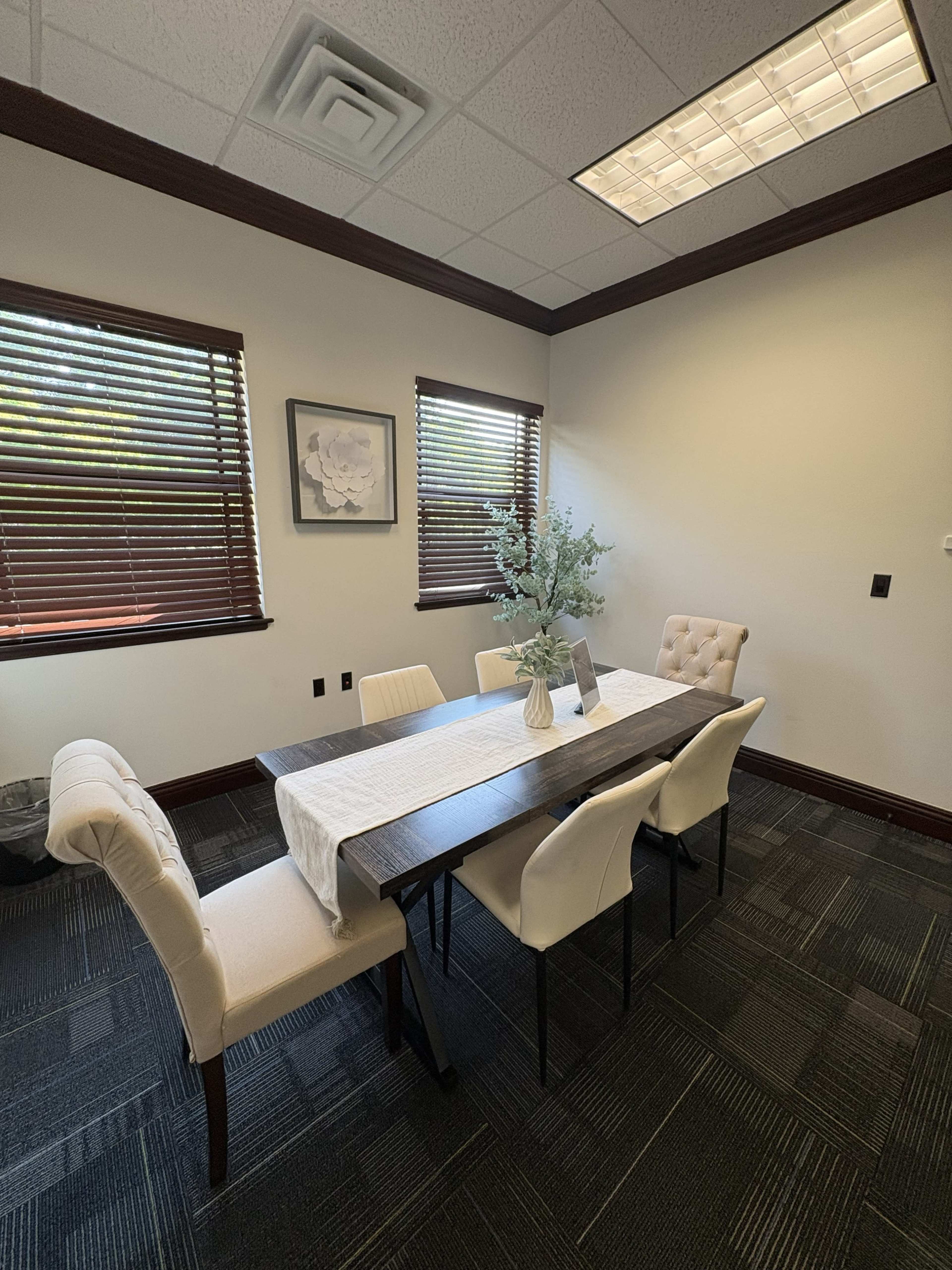 The image shows a well-lit conference room with a rectangular table, six chairs, and a decorative vase on the table, along with framed artwork on the walls.