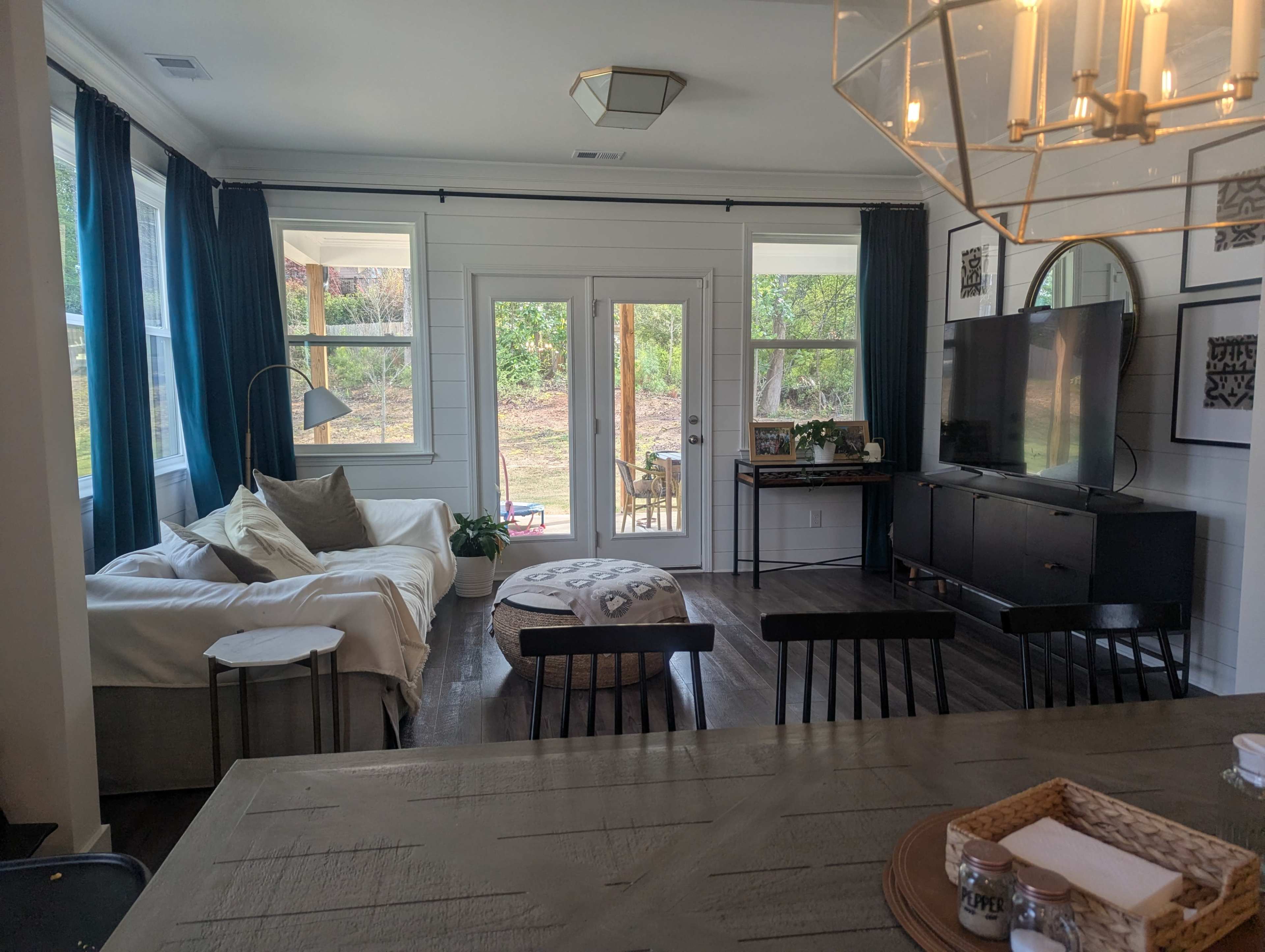 The image shows a living room with a sectional sofa, a round ottoman, a TV console, and large windows that open to a patio.