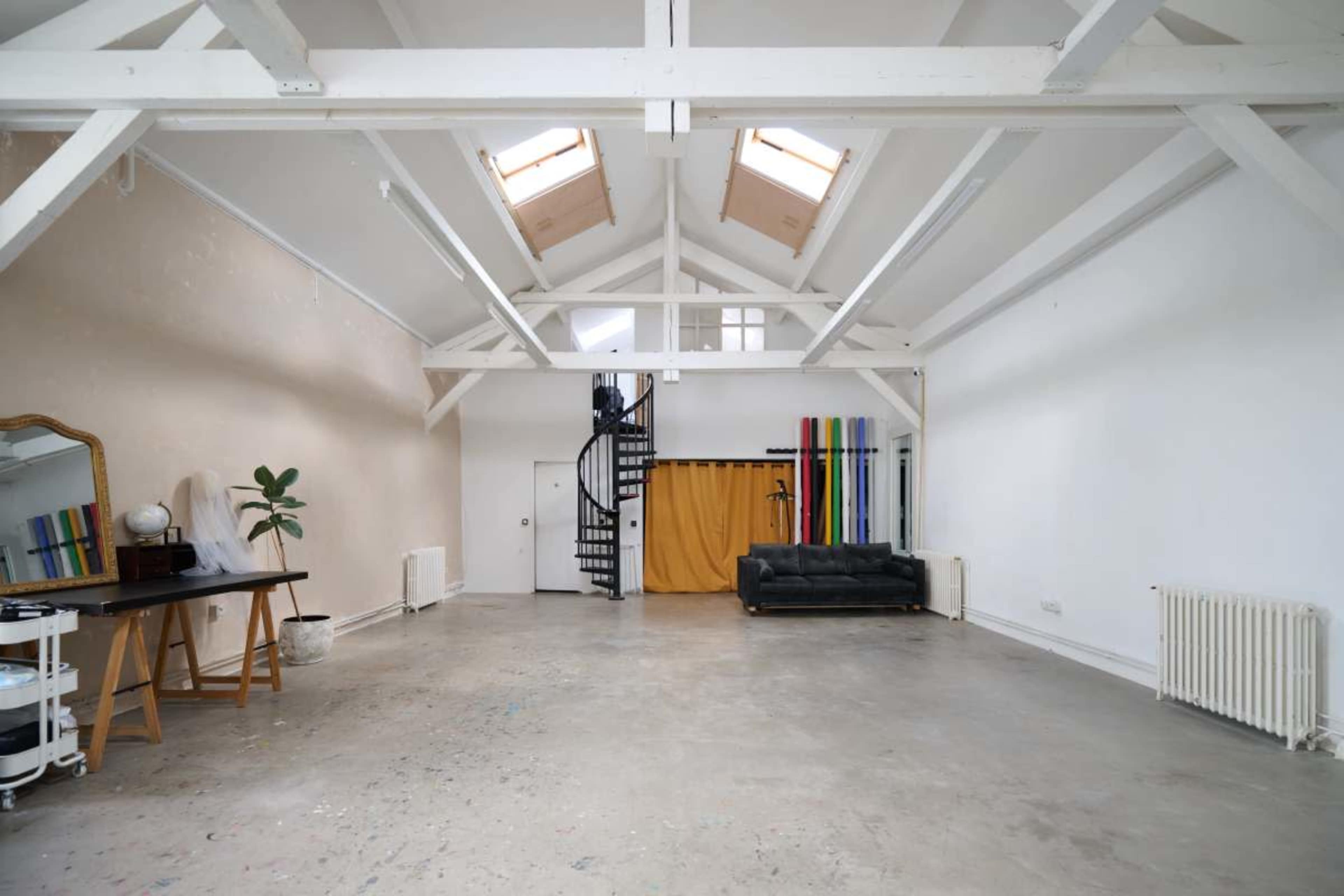 The image shows a spacious, bright loft-like interior with a wooden beam ceiling, a staircase, a black sofa, and colorful panels against one wall.