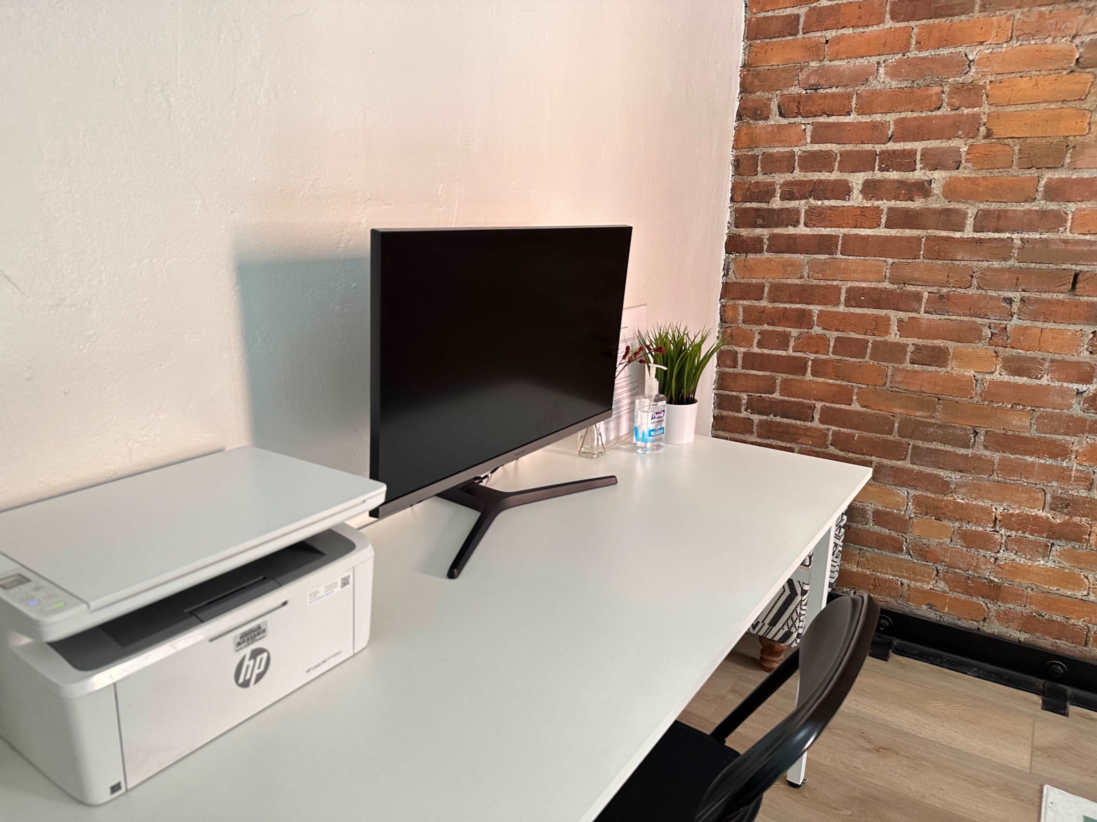A minimalistic workspace features a monitor, a printer, a water bottle, a small plant, and a chair, all set against a brick wall.