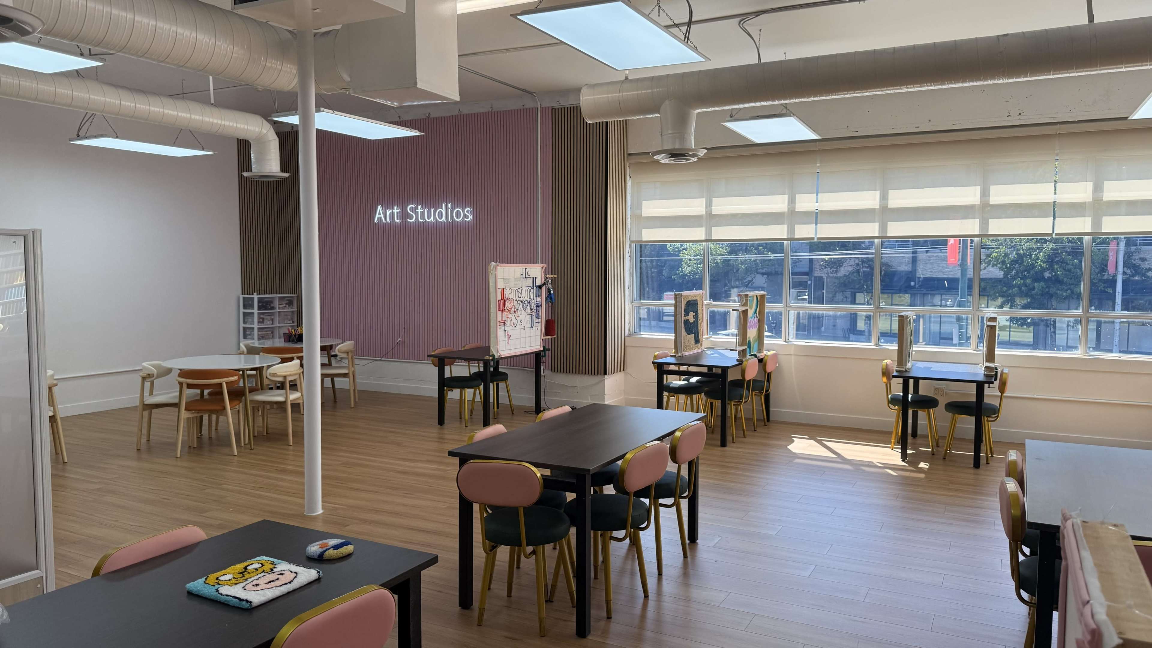 The image shows a well-lit art studio with several tables and chairs arranged throughout the space, featuring large windows that provide natural light.