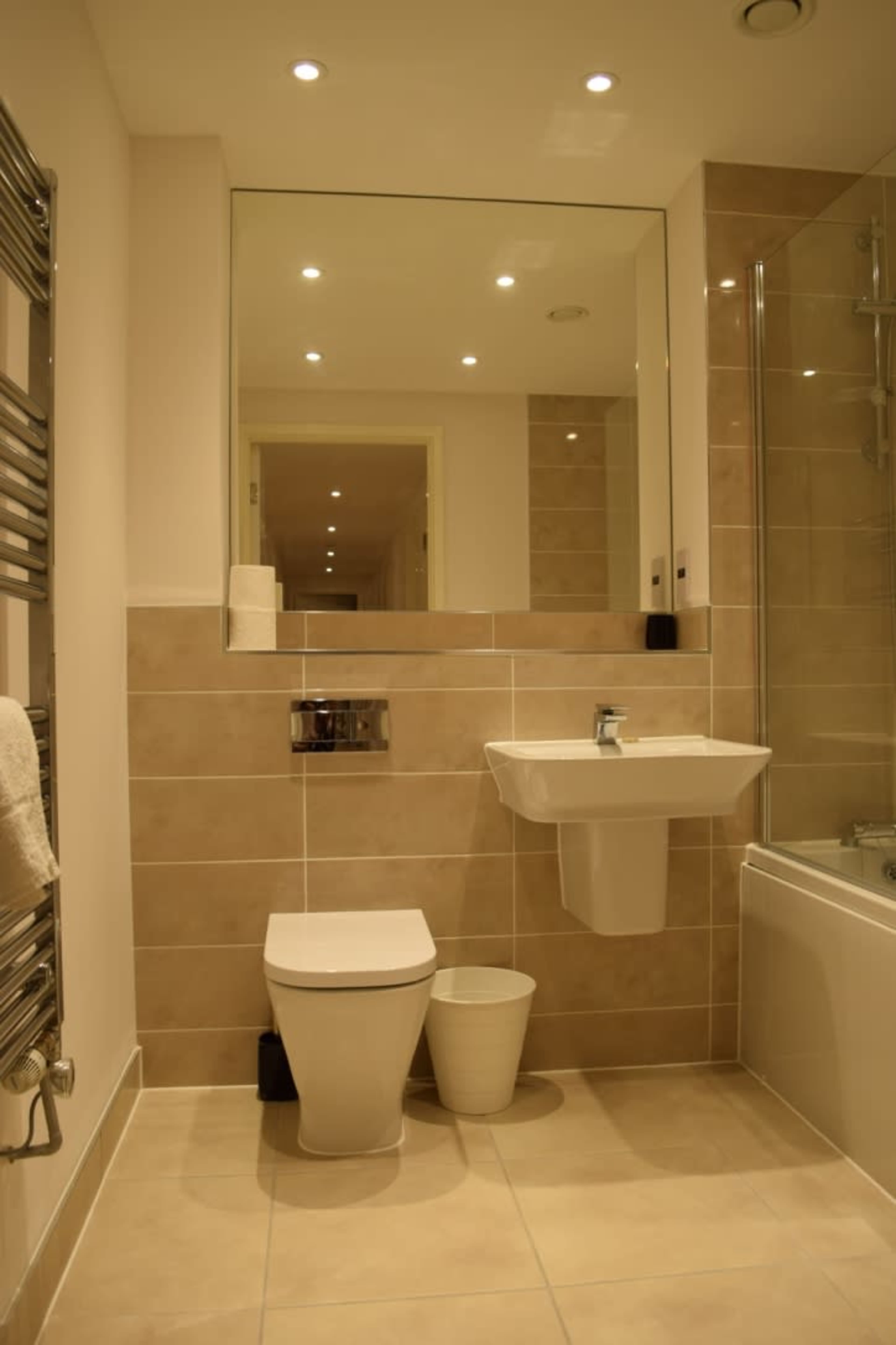 A modern bathroom featuring a wall-mounted sink, a toilet, a bathtub, and a large mirror with recessed lighting.