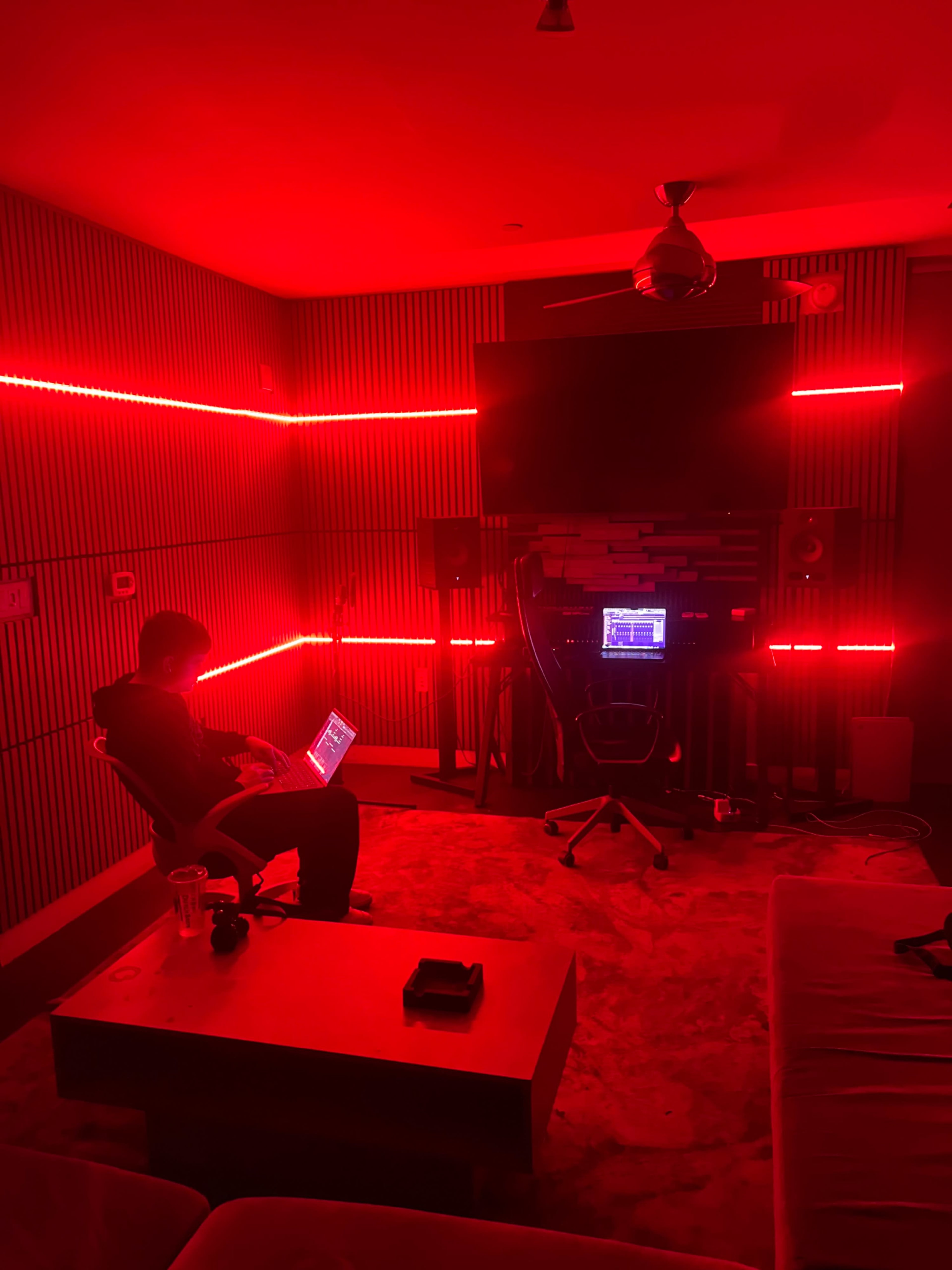 A person sits in a chair with a laptop in a room illuminated by red LED lights, surrounded by audio equipment.
