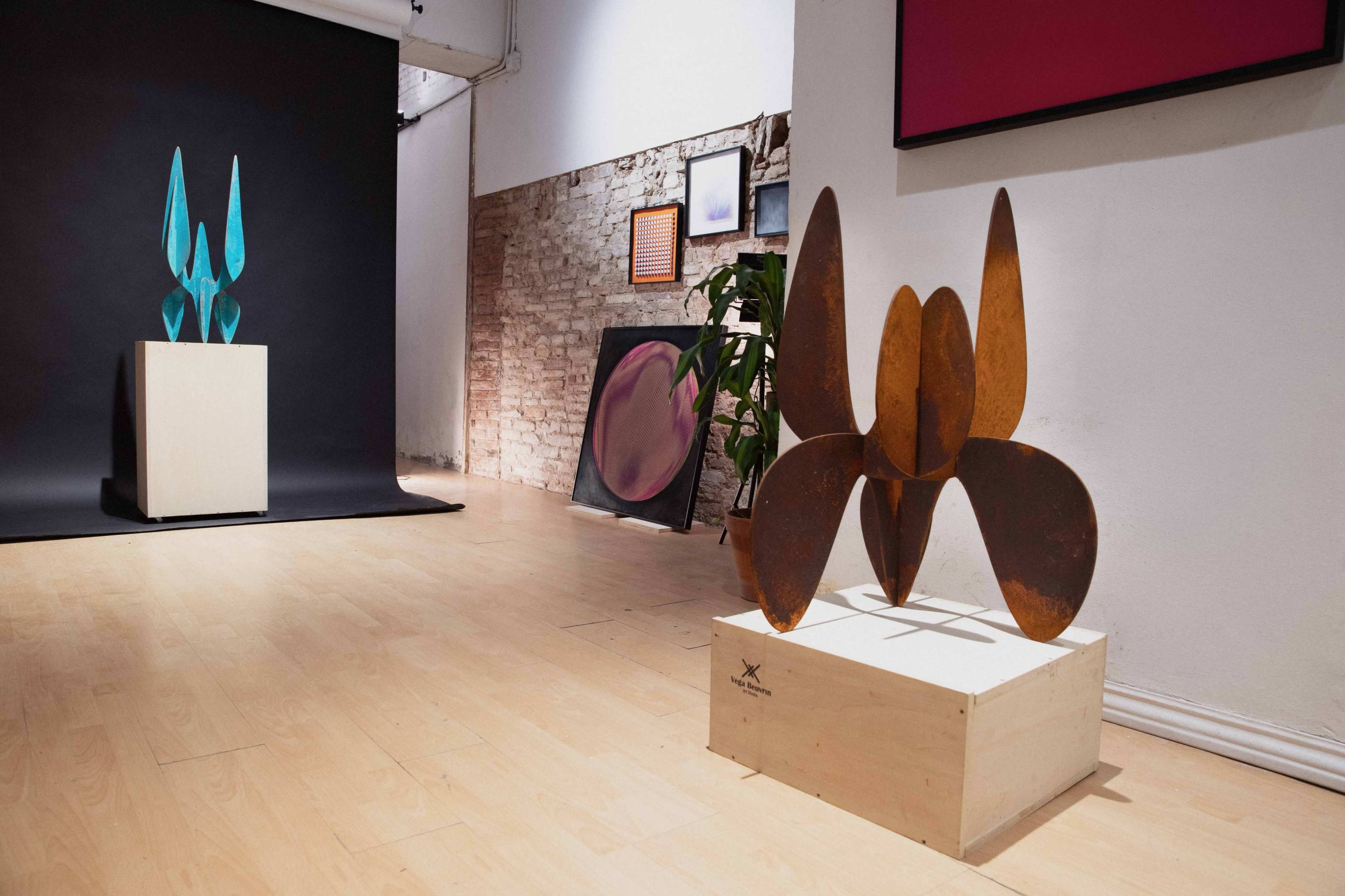 The image features an art gallery interior with two sculptures displayed on wooden pedestals, one made of blue metal and the other of rusted metal, against a backdrop of various artworks on the walls.