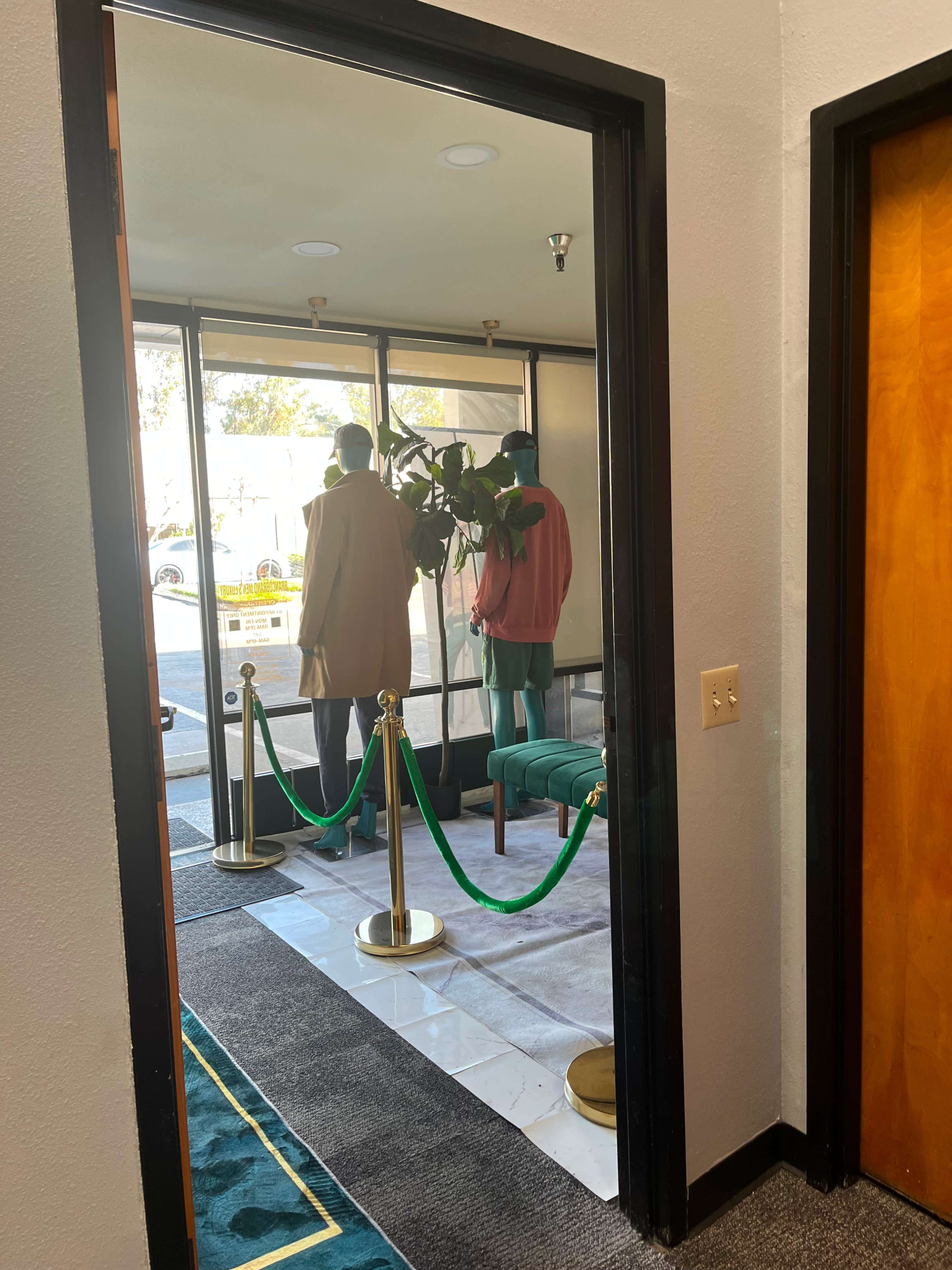 The image shows a hallway leading to a glass entrance where two mannequins are placed near a potted plant, separated by a rope barrier.