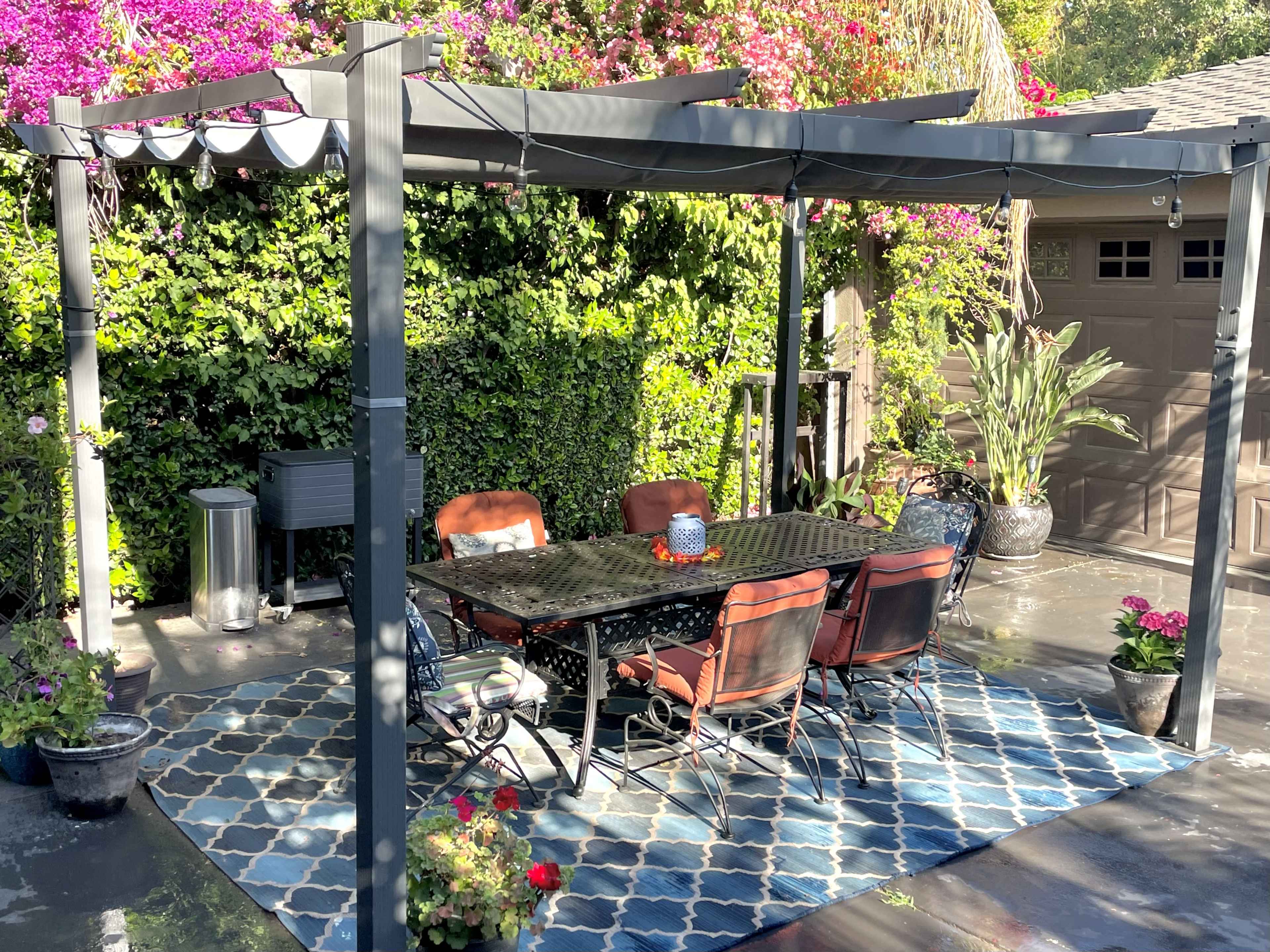 A covered outdoor dining area features a large table surrounded by chairs, with vibrant plants and flowers in the background.