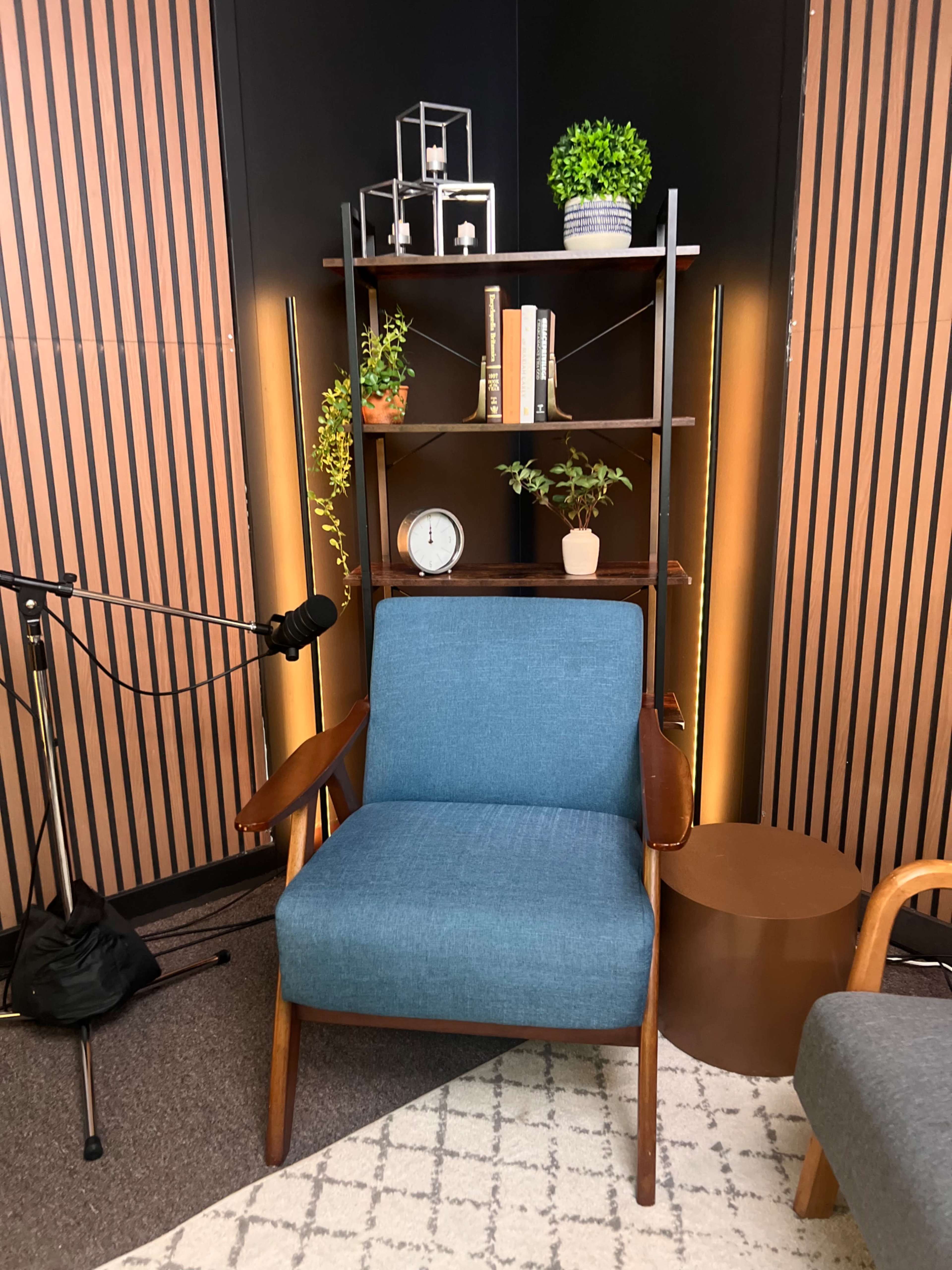 The image shows a cozy corner with a teal armchair next to a wooden bookshelf, featuring decorative plants and a clock, set against a backdrop of wooden panels and studio lighting.